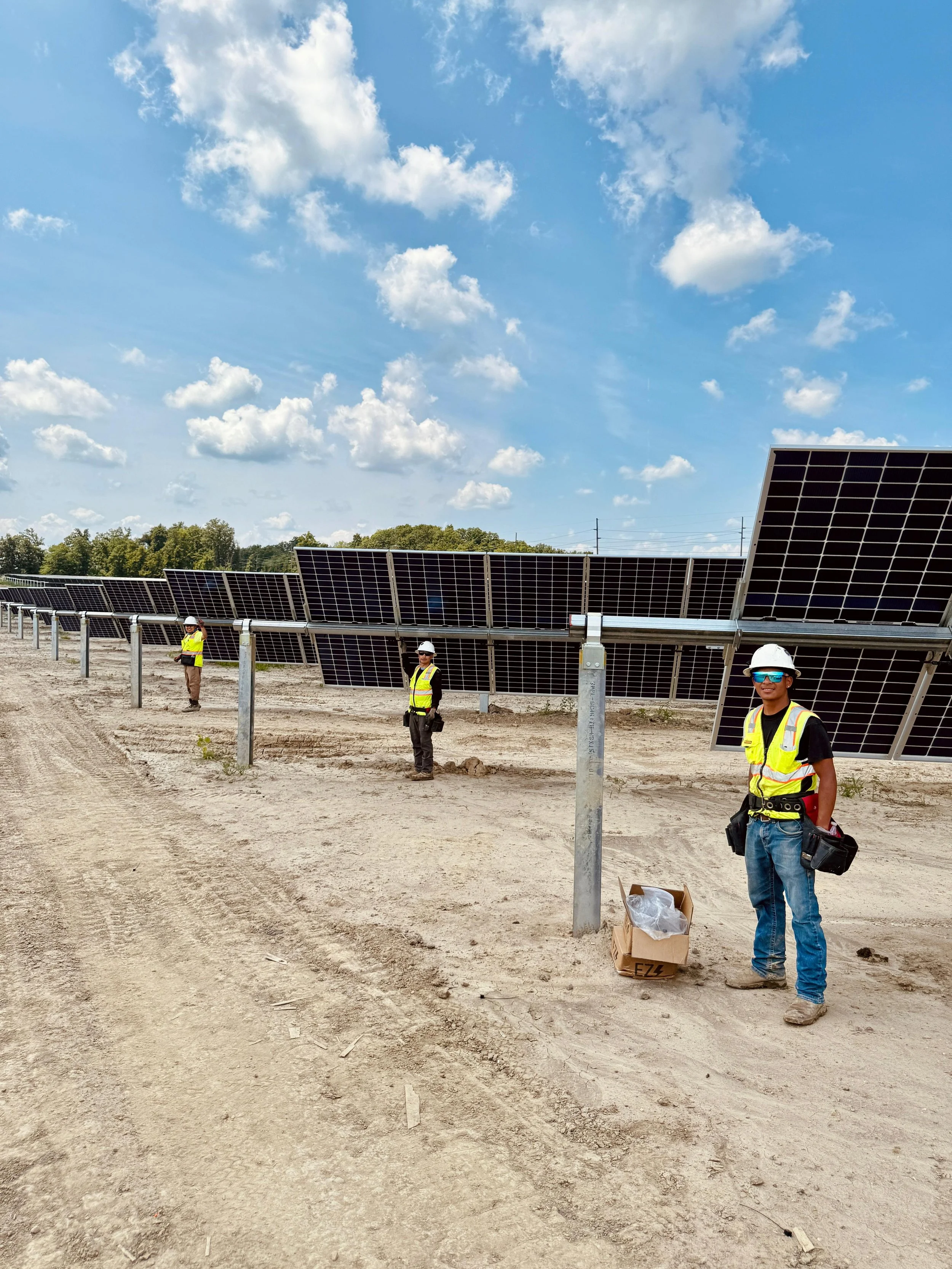 Three workers wearing safety vests and helmets installing solar panels on a barren dirt field under a partly cloudy sky.