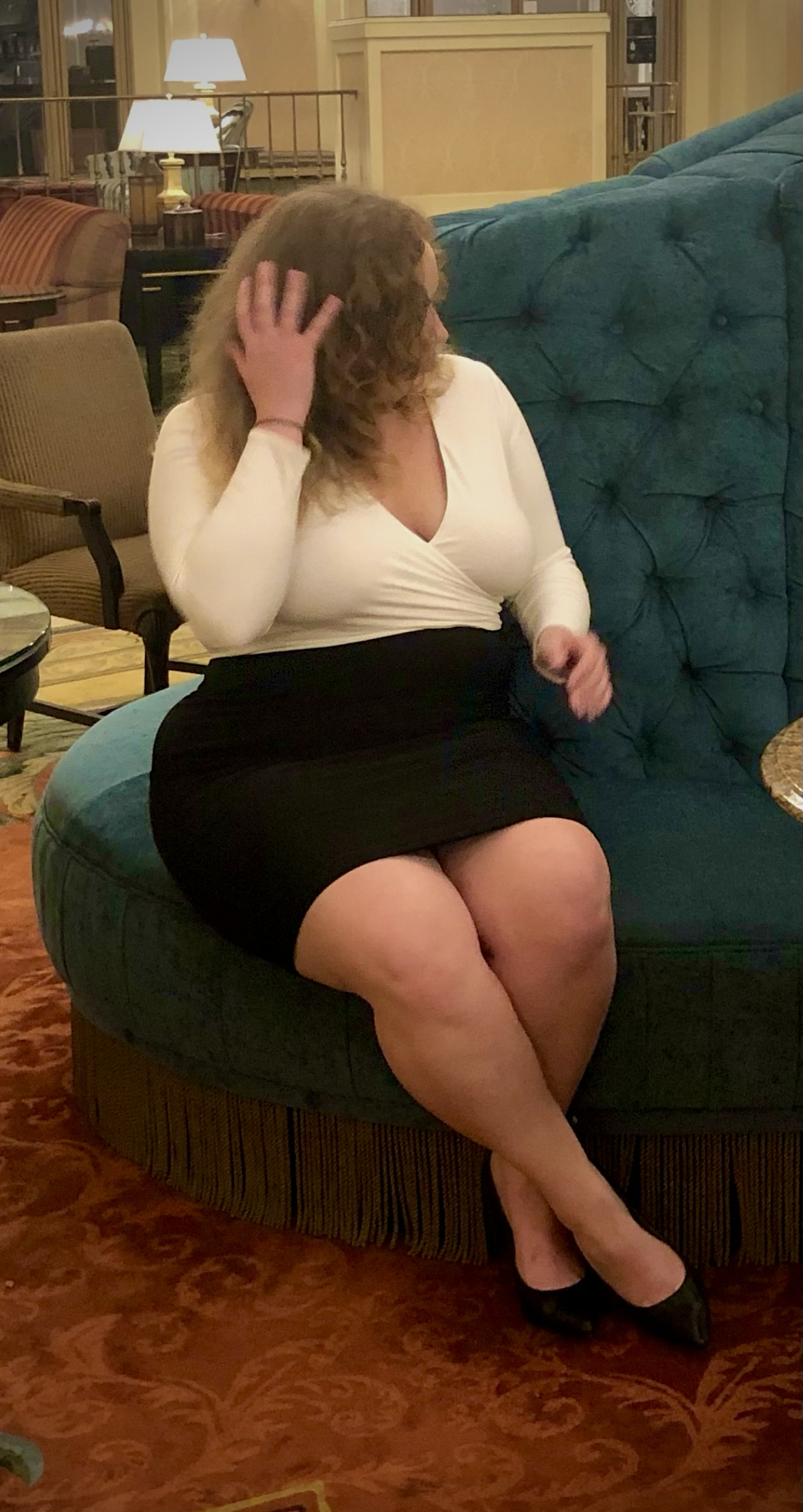 Woman with curly hair sitting on a round upholstered bench in a hotel lobby, wearing a white top, black skirt, and black heels, with her hand touching her hair.