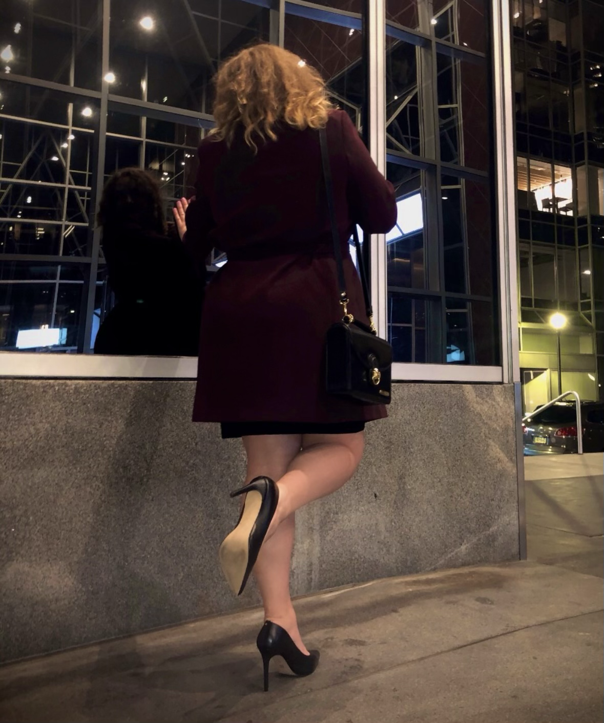 A woman with curly hair wearing a burgundy coat and black high heels standing outside at night, looking at her reflection in a glass window of a modern building.
