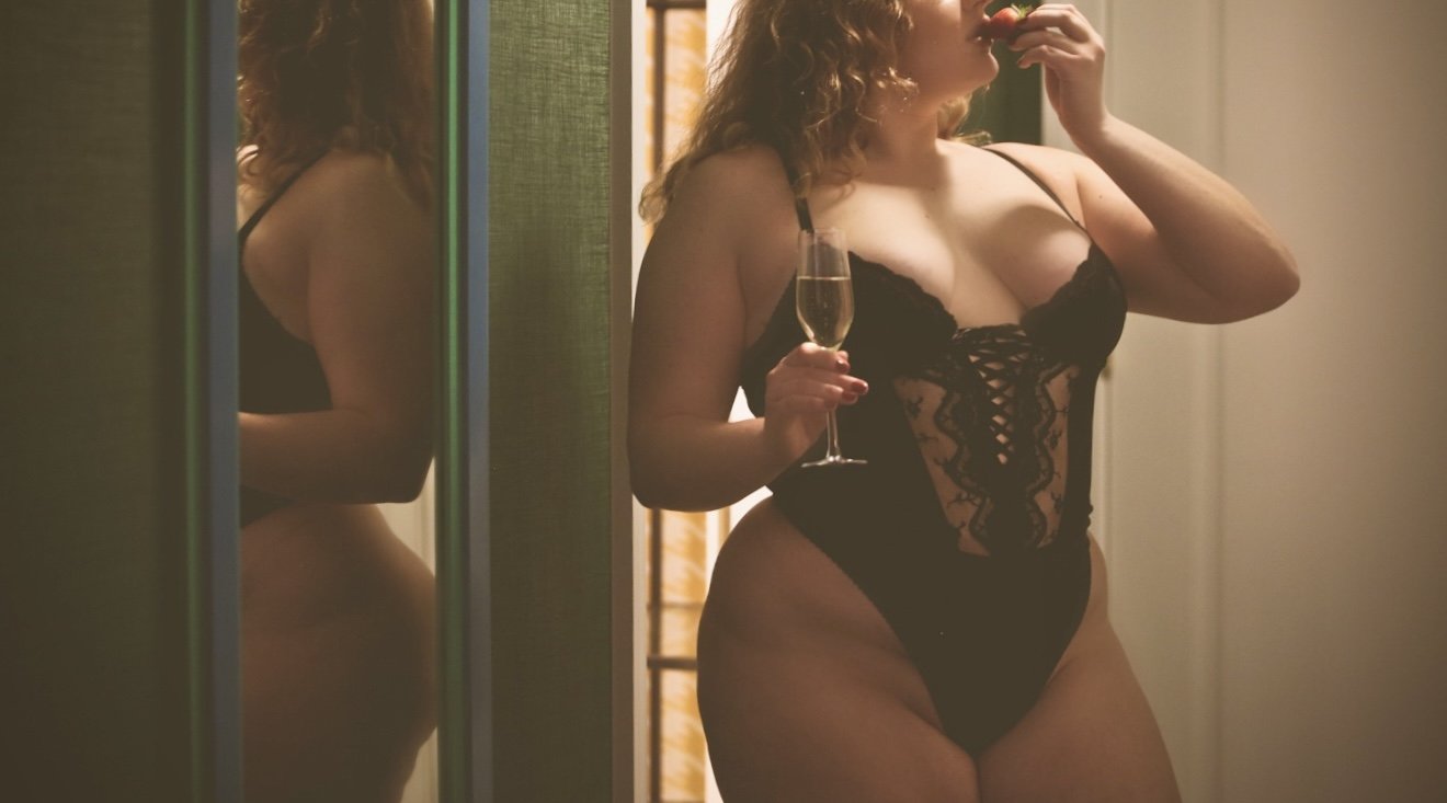 A woman with curly hair in black lace lingerie holds a glass of champagne while standing in front of a mirror.