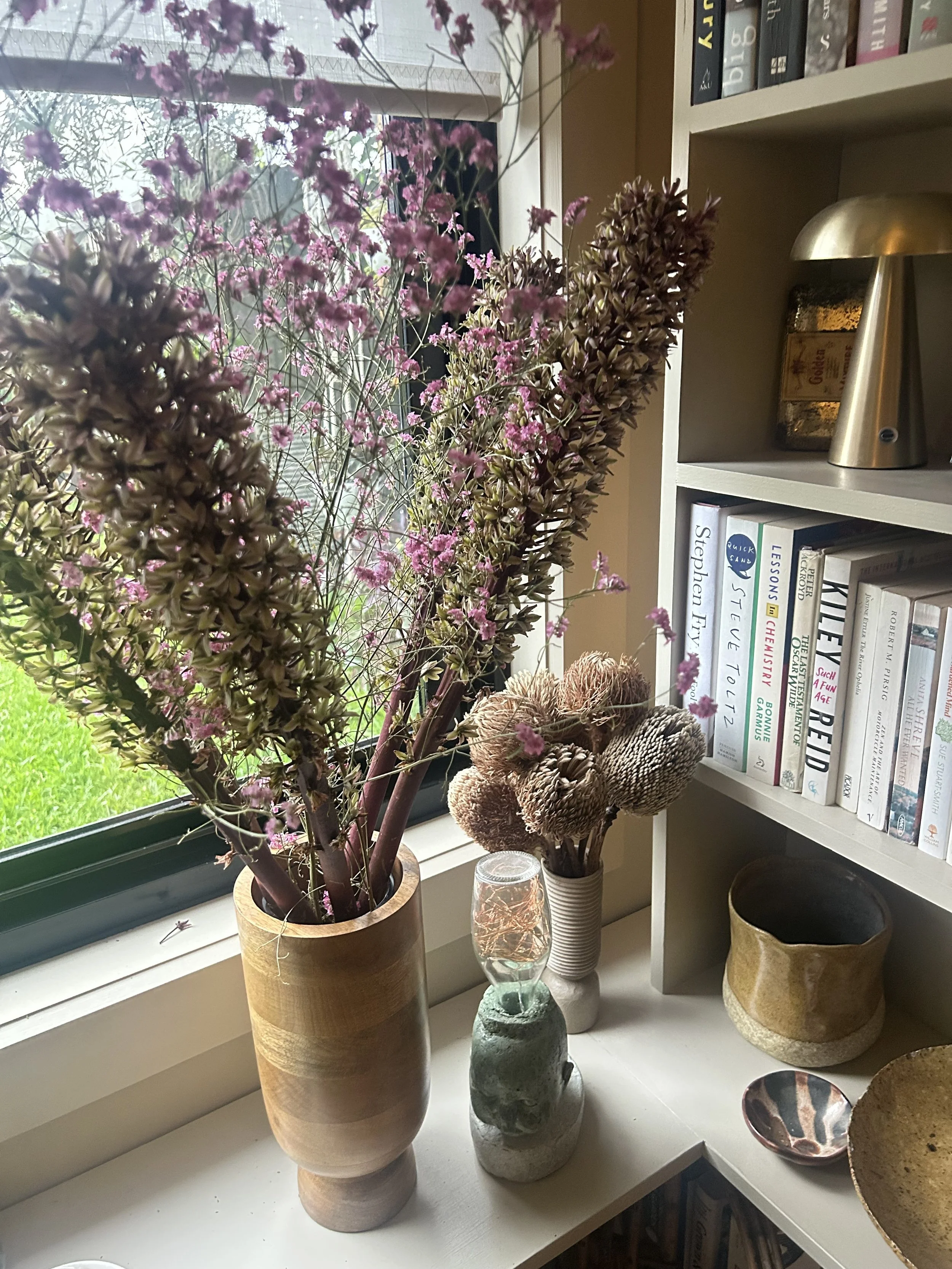 A windowsill with a large dried flower arrangement with purple and brownish foliage, a lamp made from a sculpted doll's head, a white vase with dried botanical branches, ceramic bowls and pots, and a bookshelf filled with books in the background.