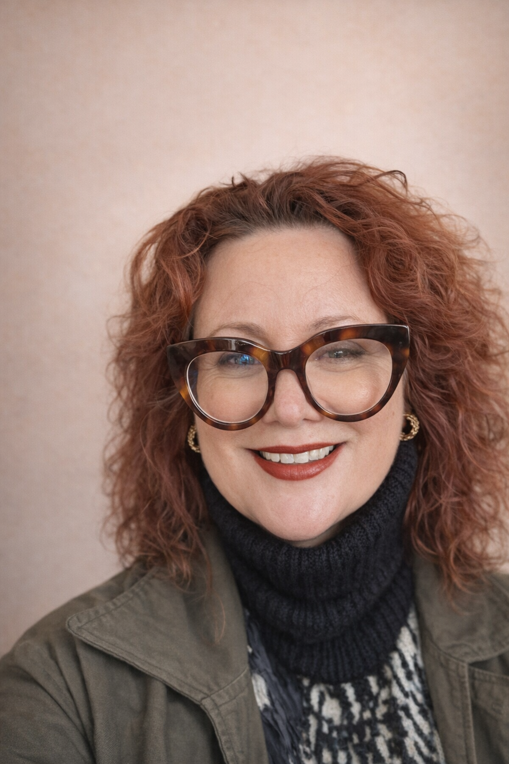 A woman with curly red hair wears large tortoiseshell glasses, gold earrings, a dark turtleneck, and a jacket, smiling at the camera against a plain background.
