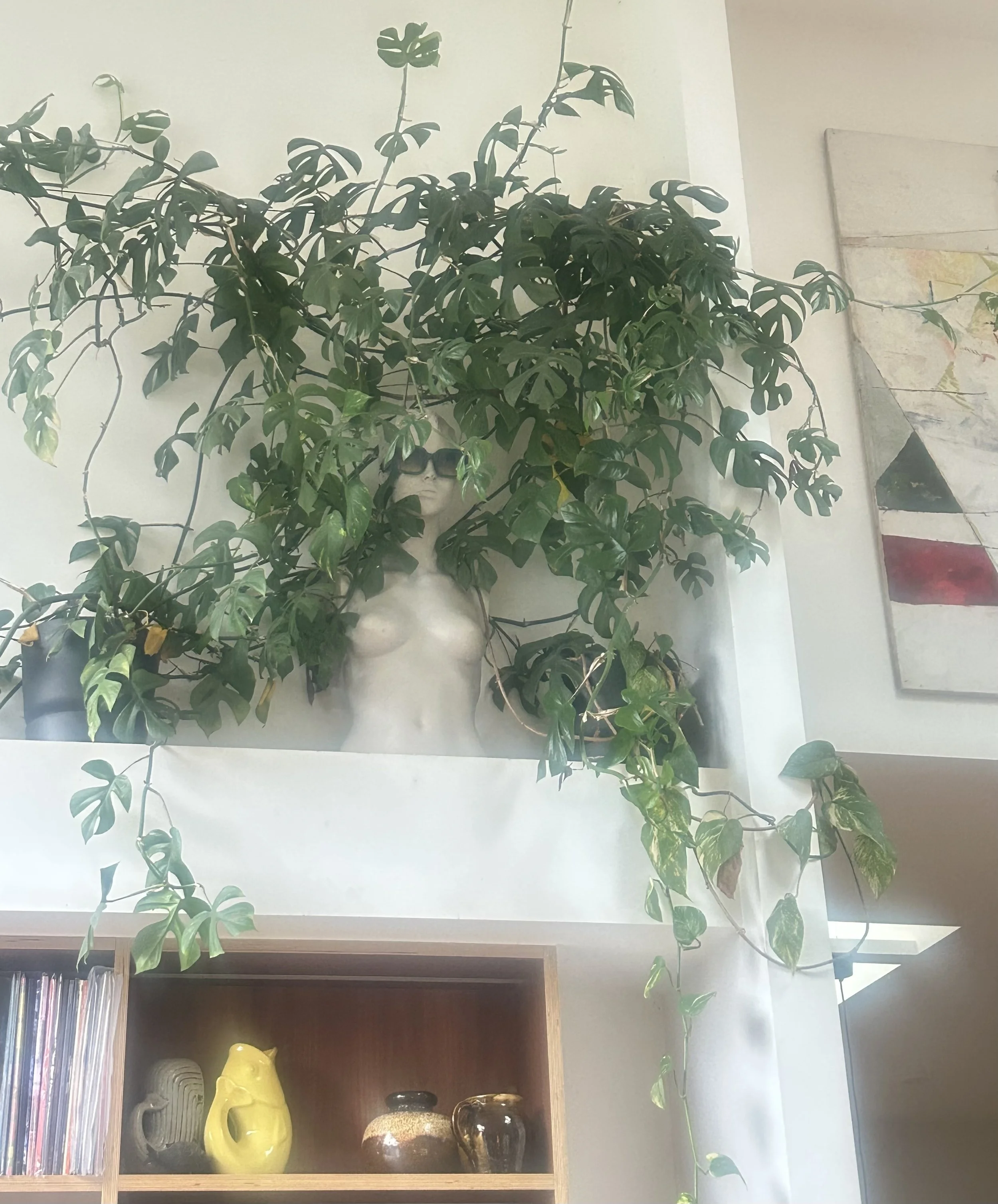 Indoor decorative scene with a monstera plant arranged in a urn-shaped vase. The plant's leaves extend above a wooden shelf that contains yellow, black, and brown patterned vases and a white ceramic piece resembling a head. To the left, a shelf with vertical stacks of magazines or records, and on the right, part of a map and an abstract painting are visible.