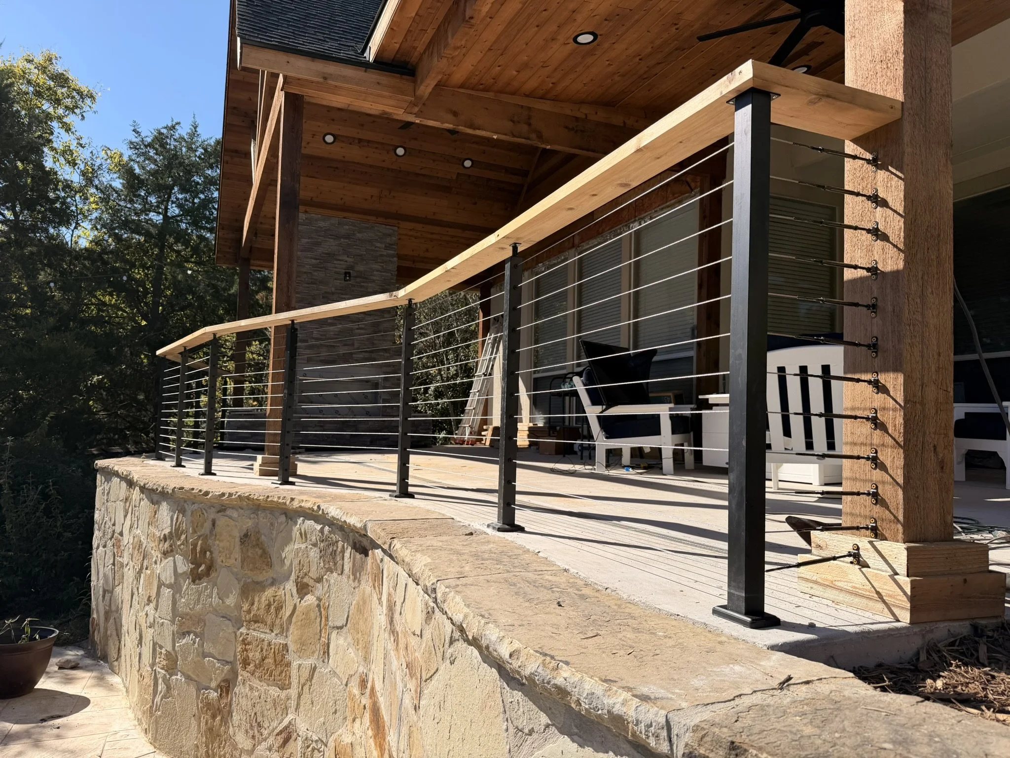 Wooden balcony with modern railing and outdoor furniture, part of a house with stone and wood exterior, surrounded by trees.