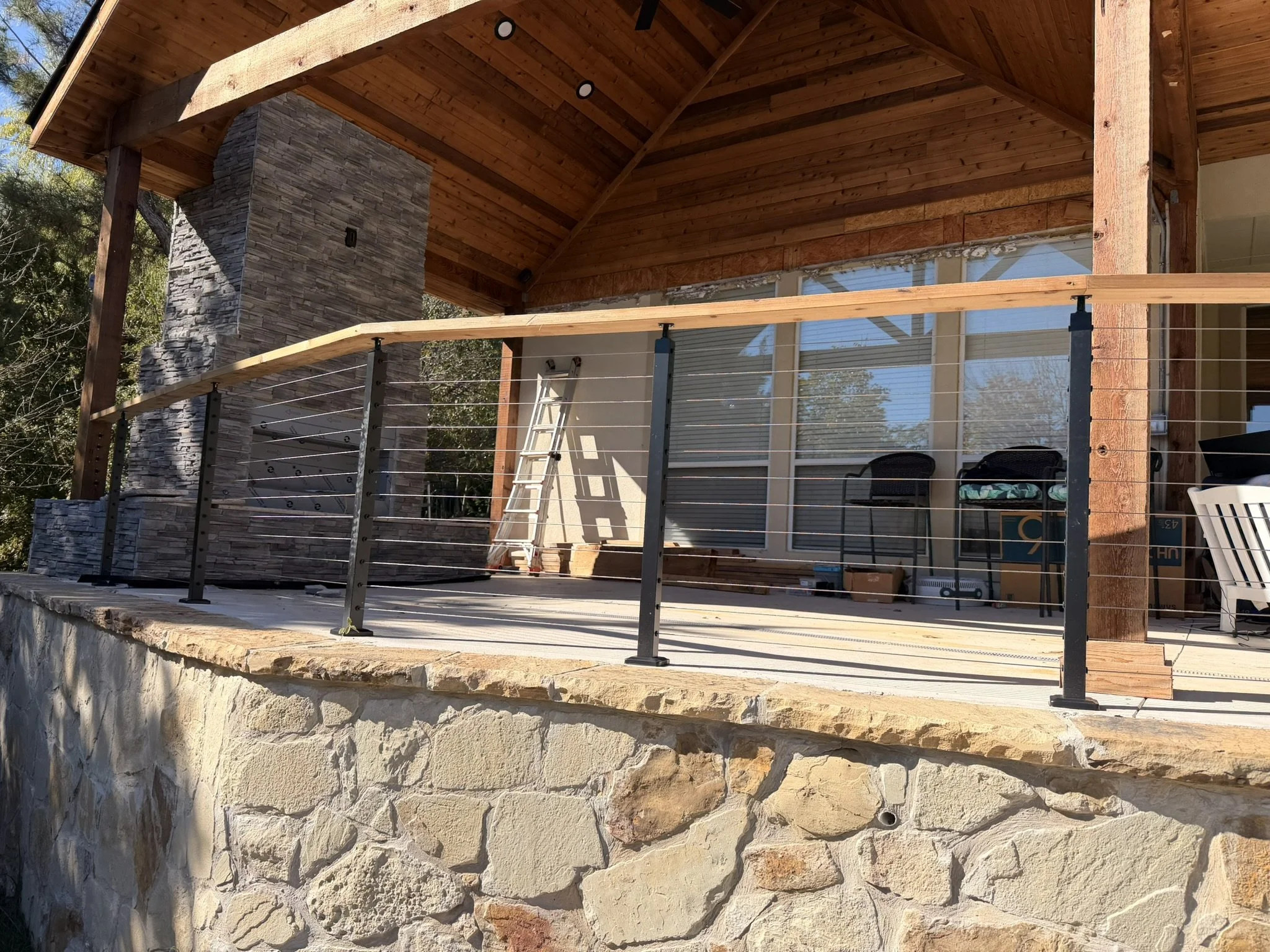 A rustic house exterior with a stone foundation and wooden porch, metal railing, ladder, chairs, and outdoor fireplace.