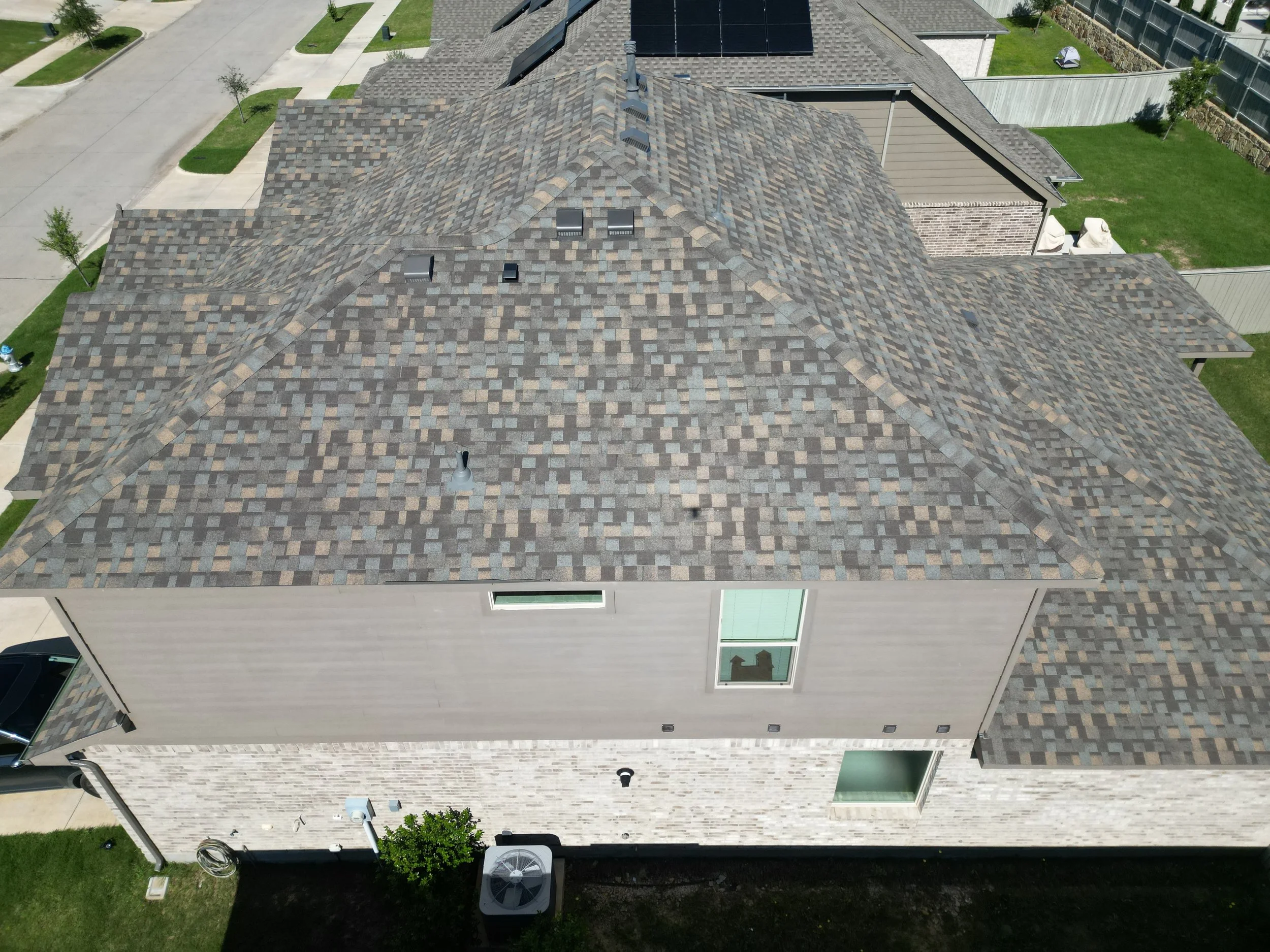 An aerial view of a house's roof with multiple sections covered in brown and gray asphalt shingles, surrounded by green grass, trees, concrete sidewalks, and neighboring houses.