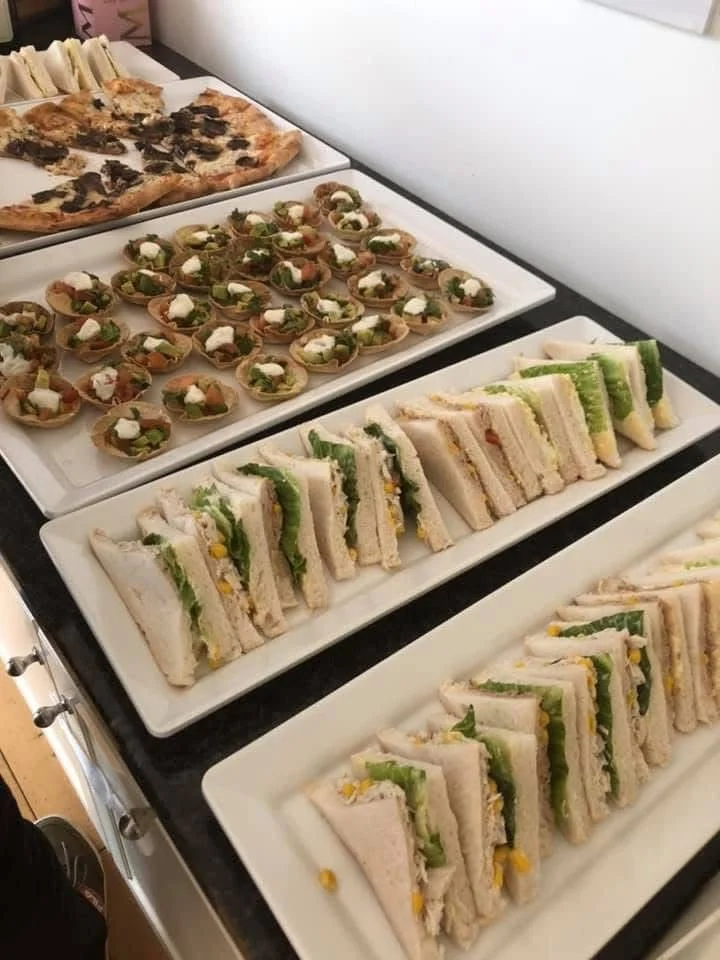 Assorted finger foods on white trays, including mini sandwiches with lettuce and egg, small open-faced tartlets with topped herbs, and slices of pizza with cheese and toppings.