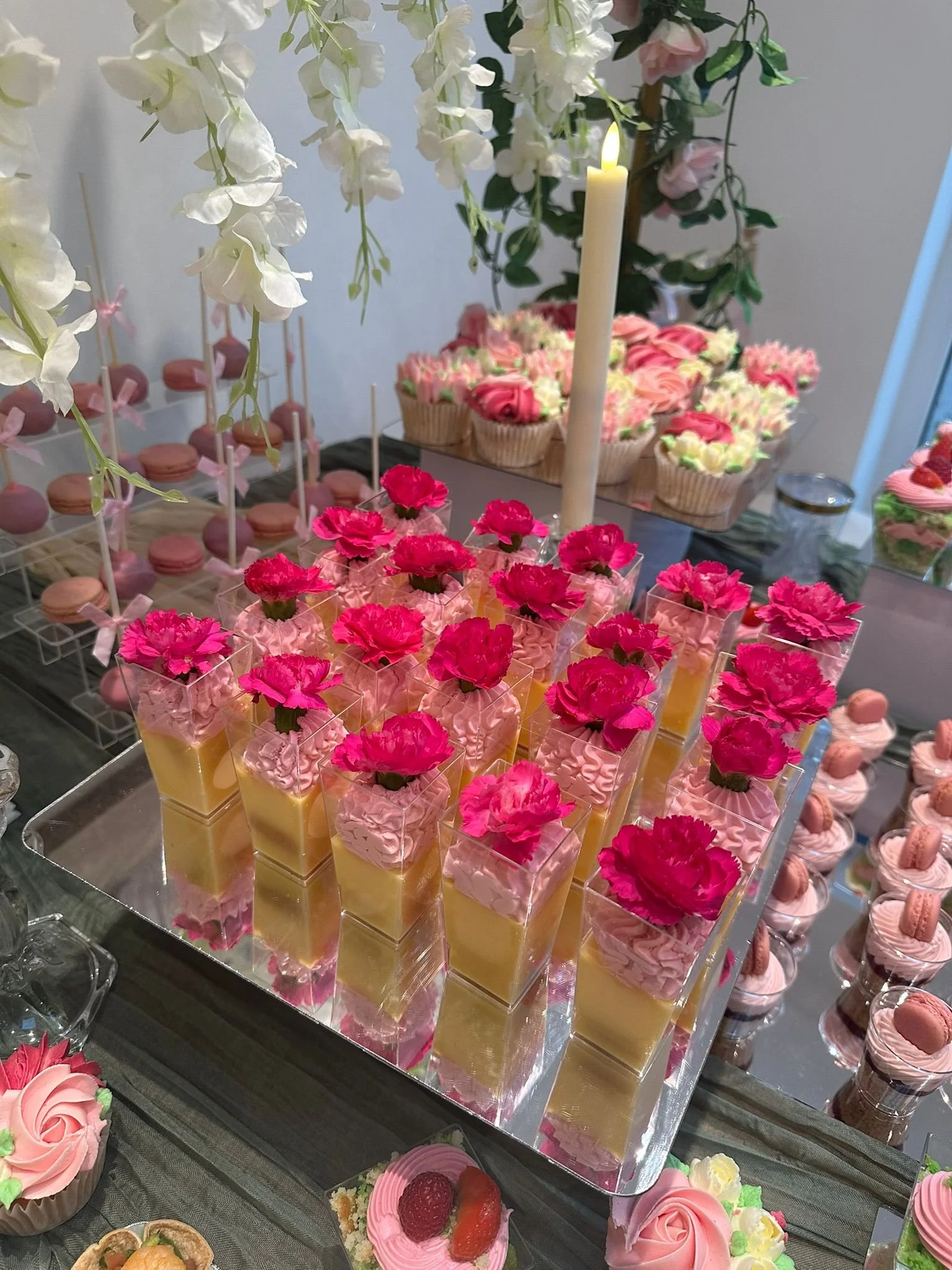 An array of pink and yellow layered desserts topped with pink flowers, displayed on a reflective silver tray, surrounded by various other pink and floral decorated confections.