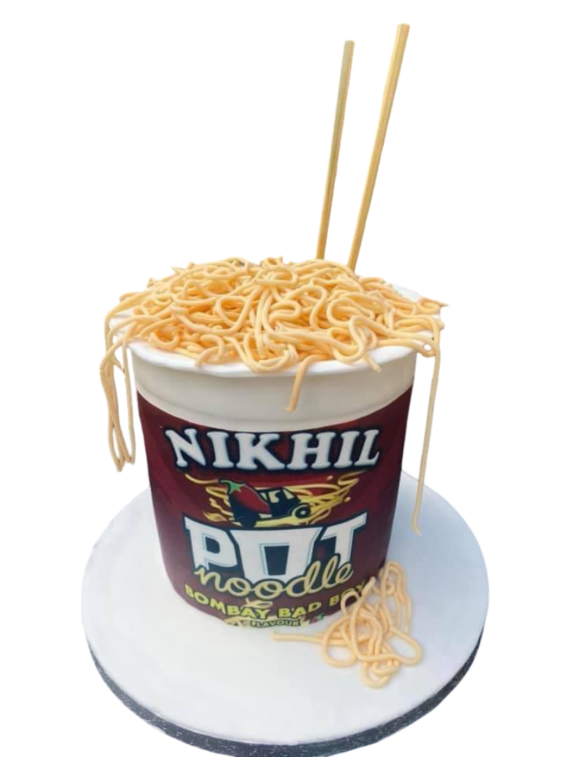 Cake designed to look like a cup of noodle soup with noodles on top and a few noodles hanging off the side, sitting on a round white platform.