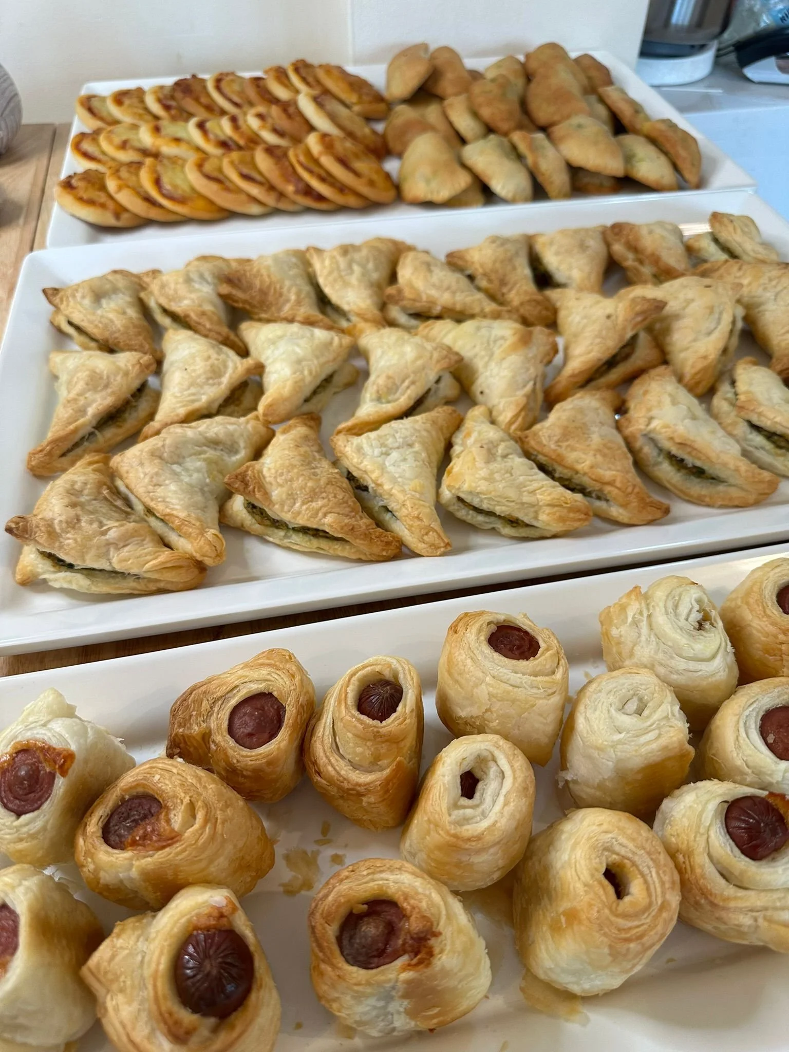 Assorted baked pastries on white platters, including hot dog pastries, samosas, triangular puff pastries, and pig-in-a-blanket rolls.