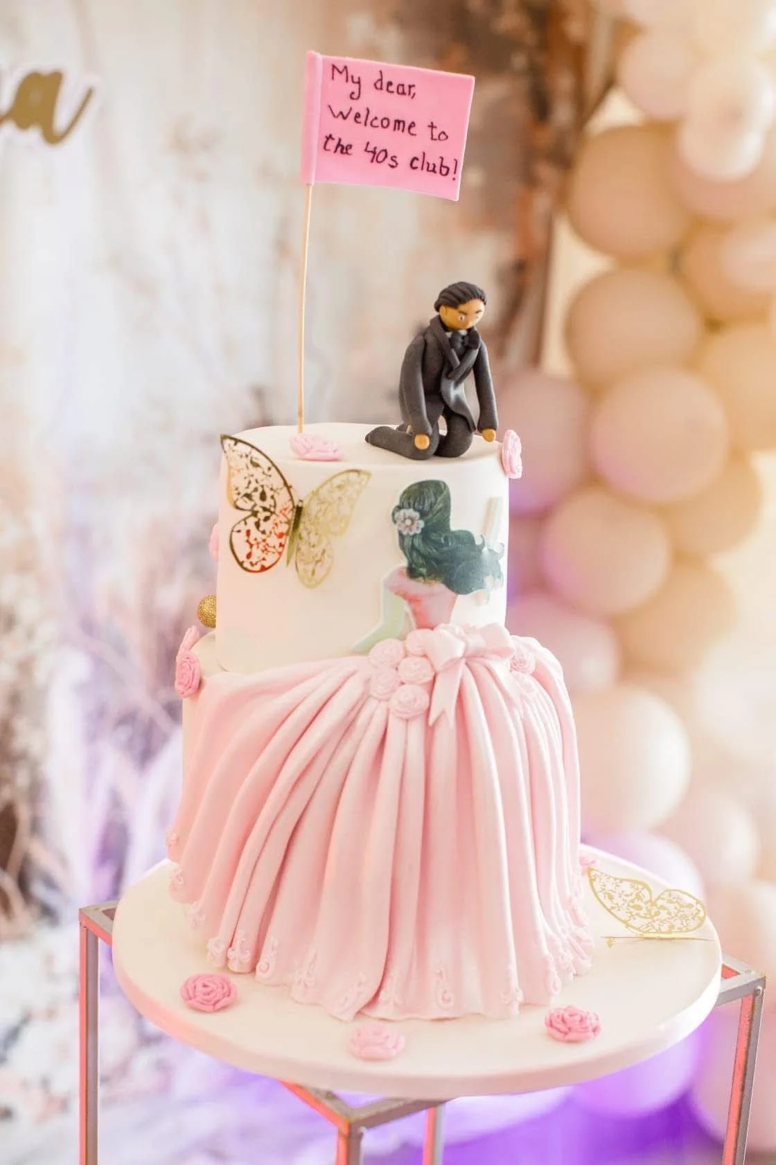 A two-tiered pink and white cake decorated with a pink dress, flowers, and butterflies, featuring a figurine of a woman kneeling in a suit on top. A pink flag reads, "My dear Welcome to the 40s club!"