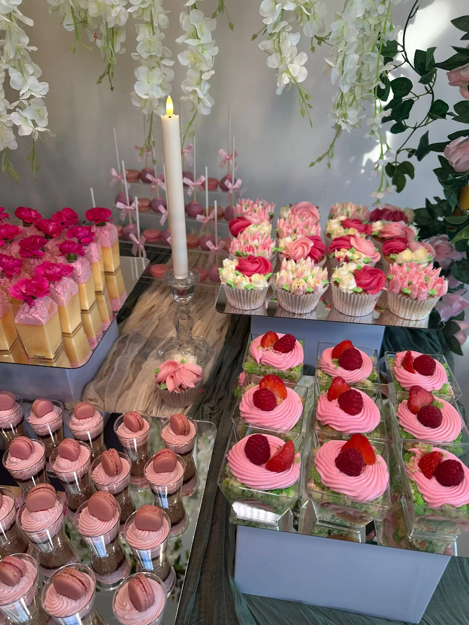 A dessert table with pink and white cupcakes topped with roses, raspberry, and strawberries, pink macarons, and other pink treats, decorated with white flowers and a lit candle.