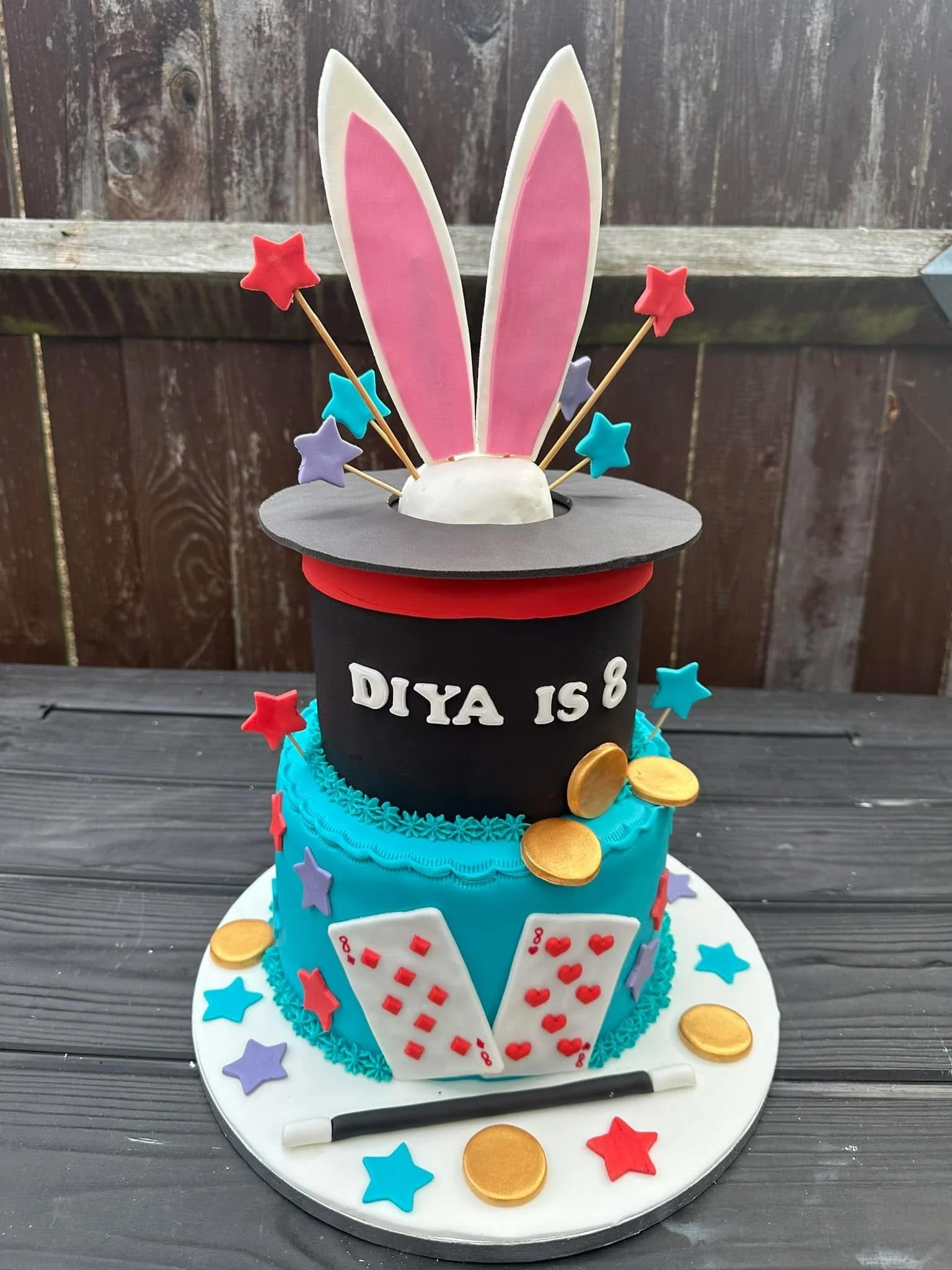 A colorful birthday cake with magician and magic theme, decorated with stars, gold coins, playing cards, and a magician's wand, with bunny ears on top and the message "DIYAS 8".