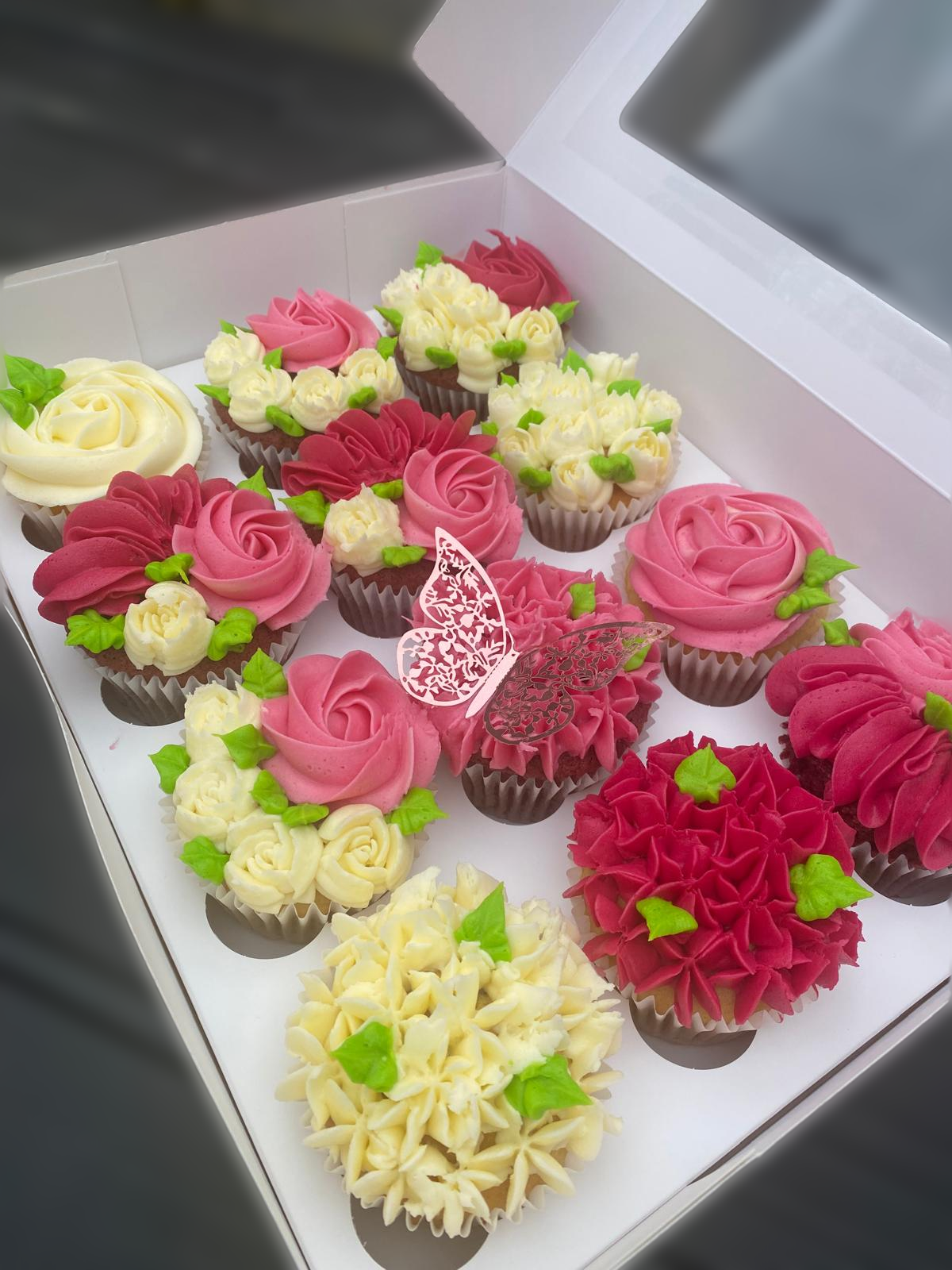 assorted cupcakes decorated with pink, white, and red icing in floral and rose designs, with green icing leaves, arranged in a white box.