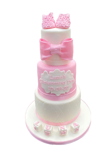 Three-tiered white and pink cake with a pink bow decoration and a plaque that reads "Luna's Christening Day 10.09.22," topped with pink shoes and blocks spelling "LUNA."