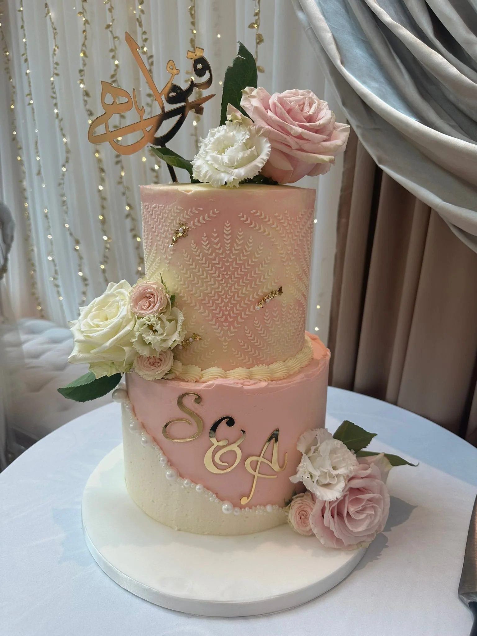 A three-tiered pink and white wedding cake decorated with pink and white roses, greenery, gold accents, and a topper with the names "S&E" and an initial. The cake is placed on a white cake board on a table with a white tablecloth.