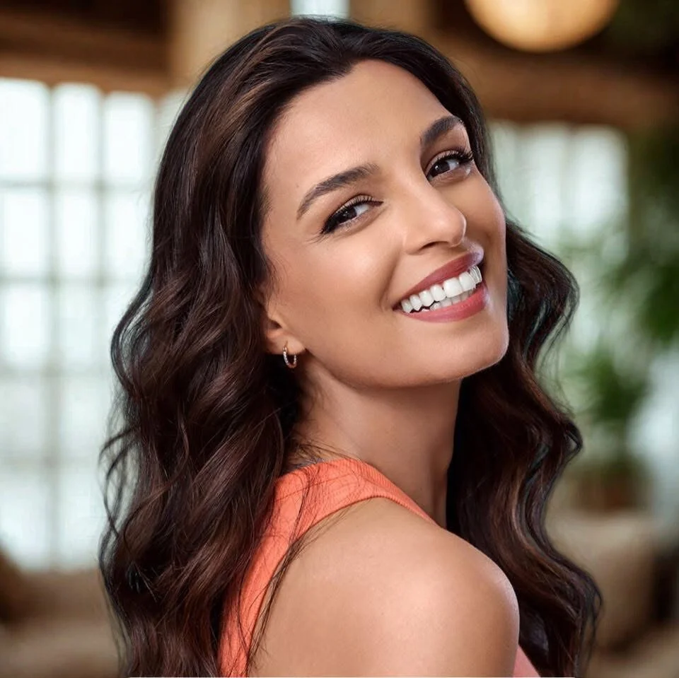 A woman with long dark wavy hair smiling, wearing an orange top, indoors with a blurred background of windows and plants.