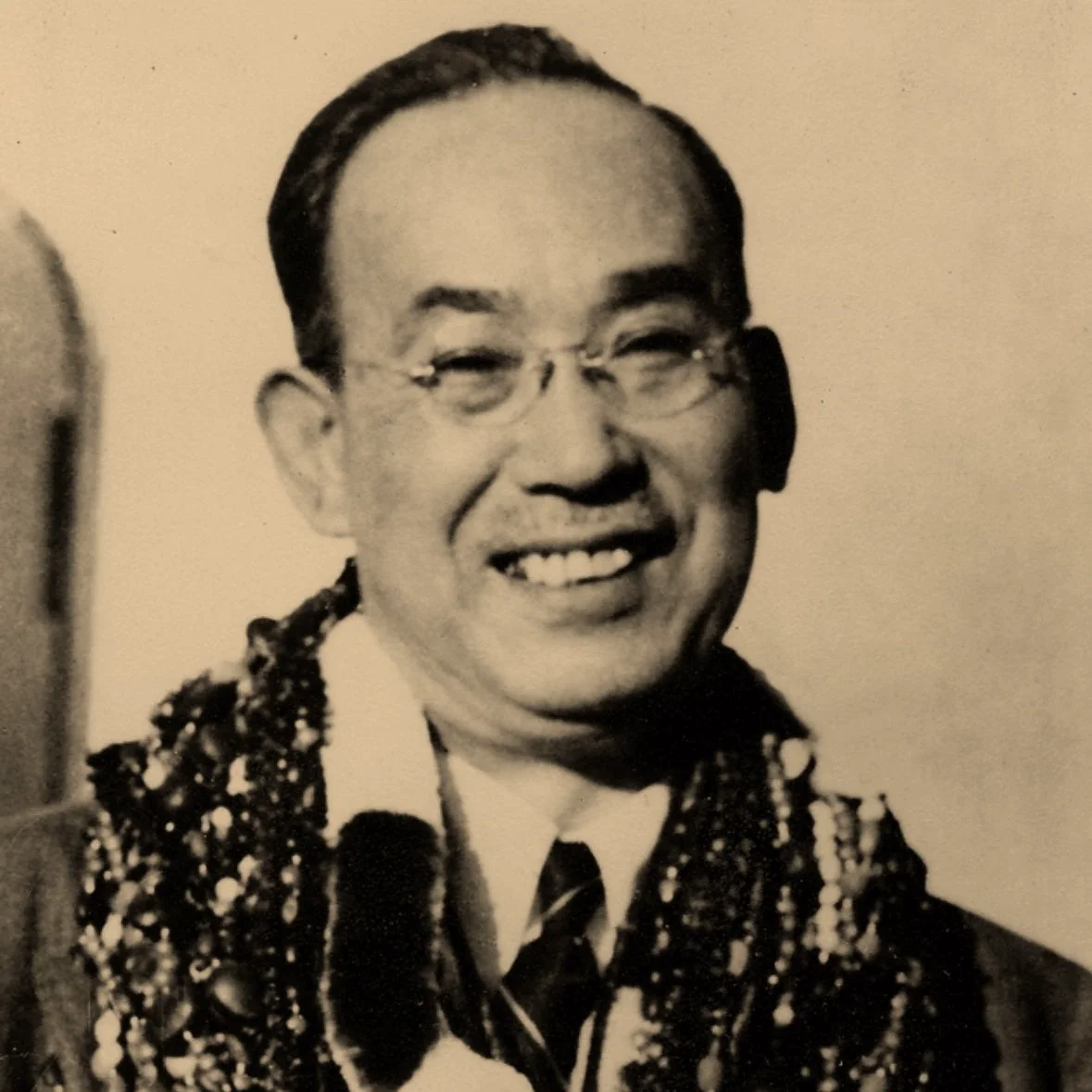 Black and white photo of a smiling man wearing glasses, a tie, and a beaded necklace.