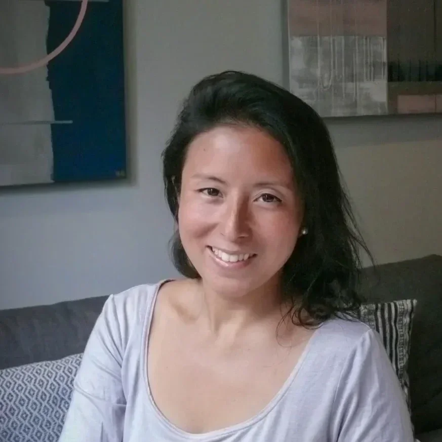 A woman with black shoulder-length hair smiling while sitting on a sofa in a living room.