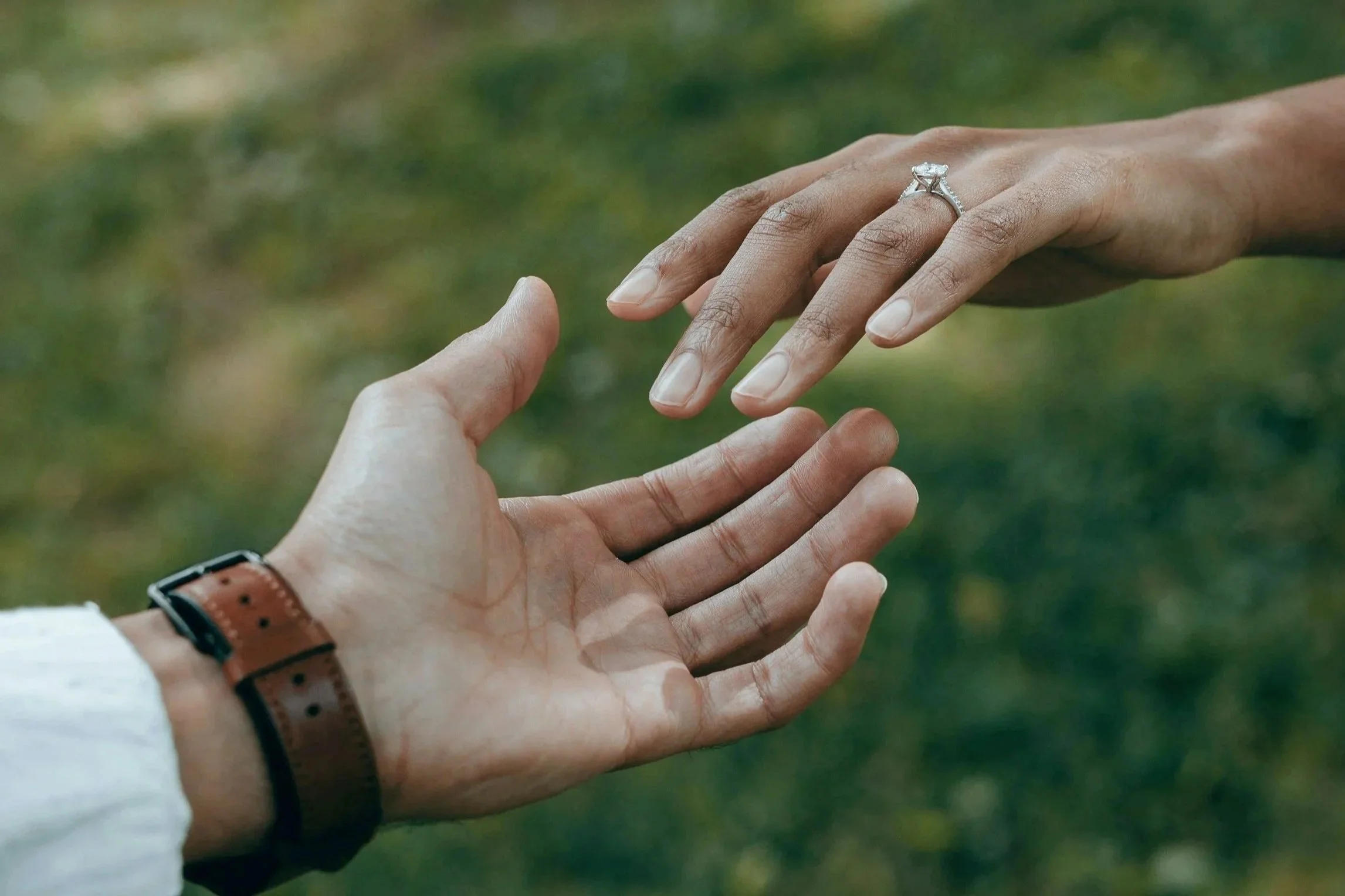 Close-up of a person’s hand reaching out to another person's hand, with a diamond engagement ring on the finger, outdoors with green grass in the background.