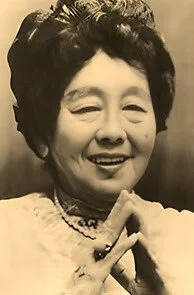 Black and white photo of an elderly woman smiling, with her hands clasped in front of her chest.