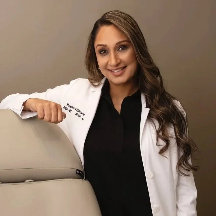A woman with long brown hair, smiling, dressed in a medical coat, leaning on a piece of medical equipment, in a clinical setting.