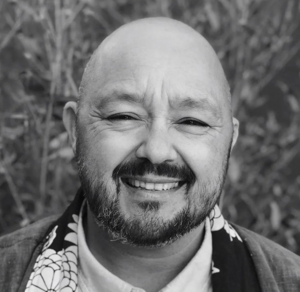 A smiling man with a beard and bald head, wearing a dark jacket and a patterned scarf, outdoors with blurred foliage in the background.