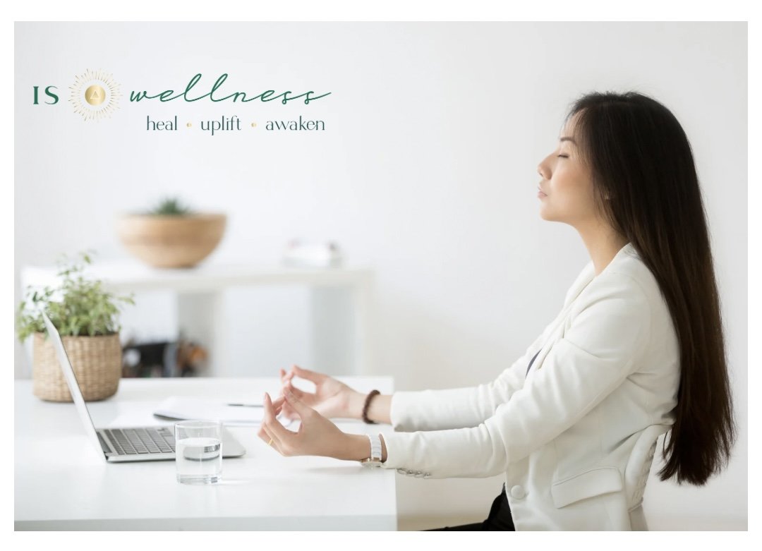 A woman practicing meditation at her desk in a bright, minimalist office with a laptop, a glass of water, and potted plants, promoting wellness and relaxation.