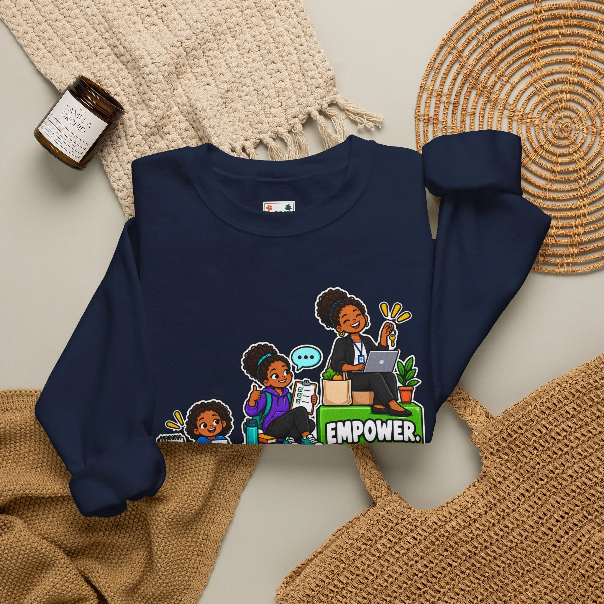 Flat lay of a navy blue sweatshirt with a graphic of diverse women working and the words "Teach" and "empower" on it, a beige textured scarf, a brown candle labeled "vanilla orchid," a round woven rattan placemat, and a woven straw hat.