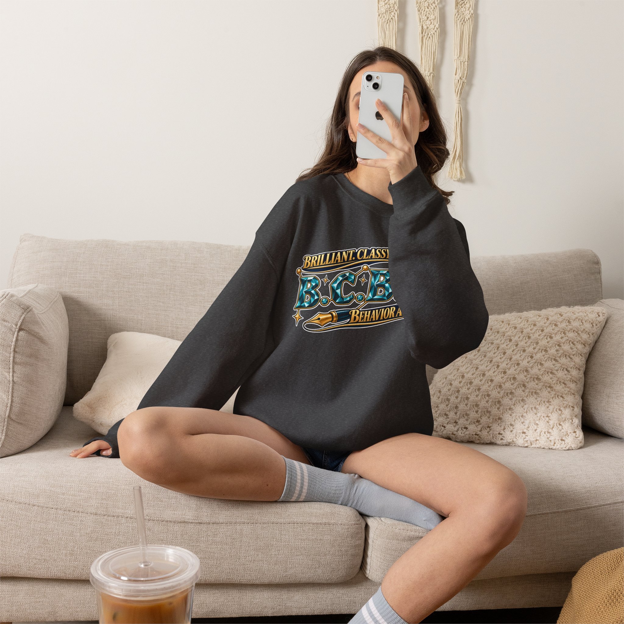 A woman with long dark hair is sitting on a light-colored couch, taking a selfie with her smartphone. She is wearing a black sweatshirt with colorful text and graphic designs. In front of her, on the table, there's a plastic cup with a straw containing a brown iced beverage.