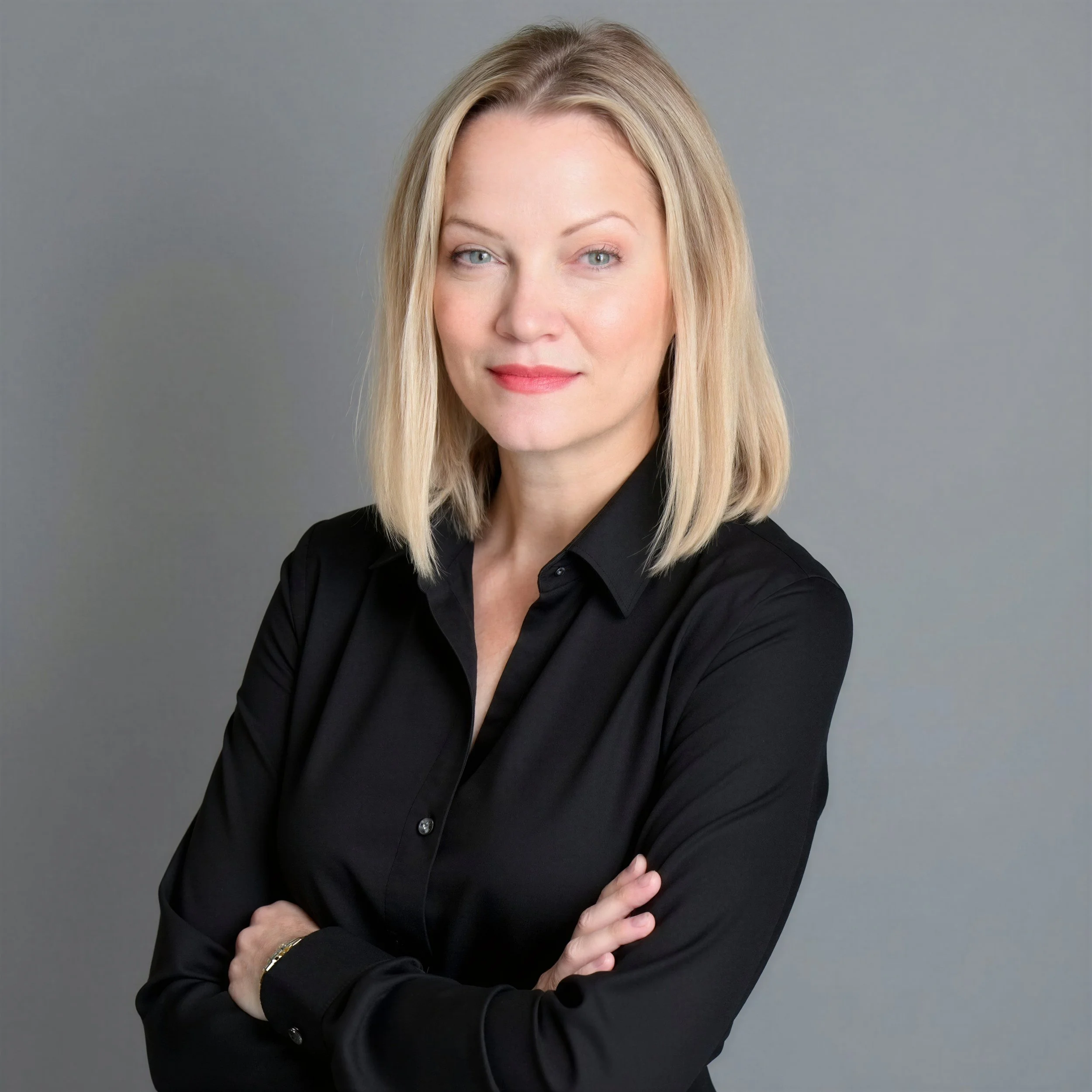 Close-up portrait of a woman with blonde hair, blue eyes, and a slight smile, wearing a black shirt and standing against a gray background.