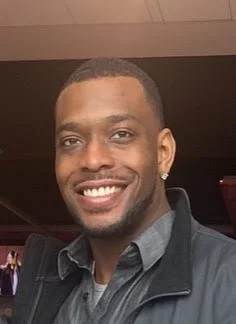 A smiling man with short hair, wearing a dark jacket and a collared shirt, in an indoor setting.