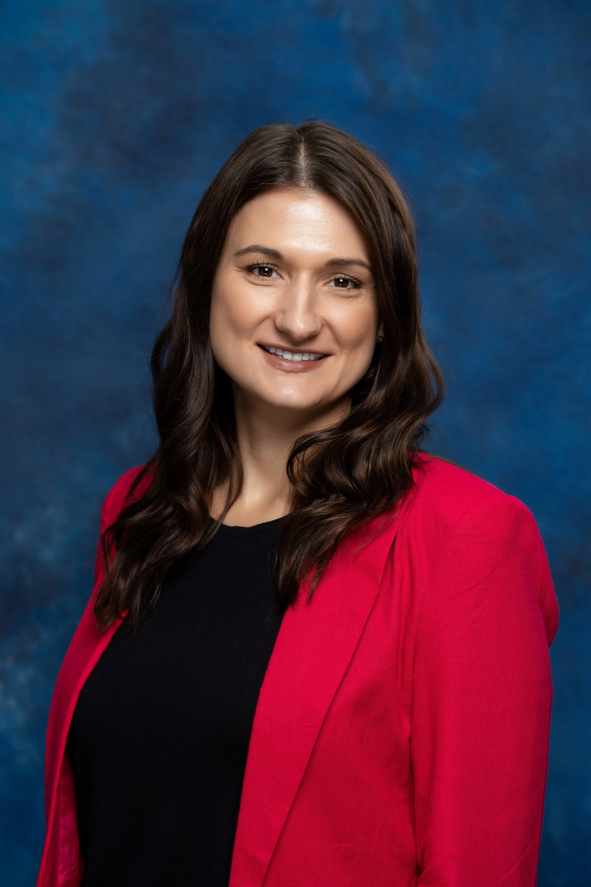 A woman with long, wavy brown hair, wearing a red blazer over a black top, smiling against a blue textured background.