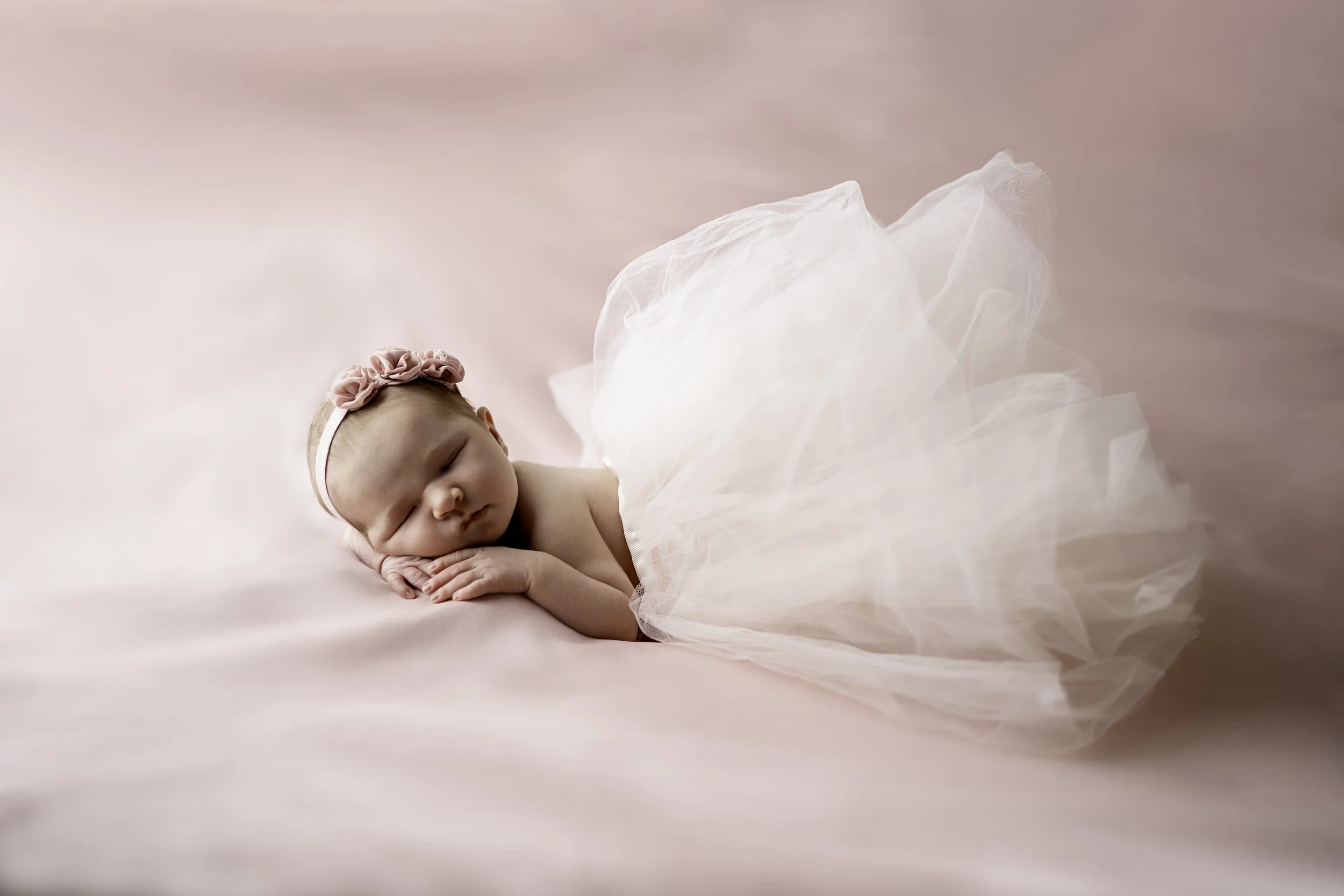 A sleeping baby girl lying on a soft pink surface, wearing a pink headband with a flower and a fluffy white tulle skirt.