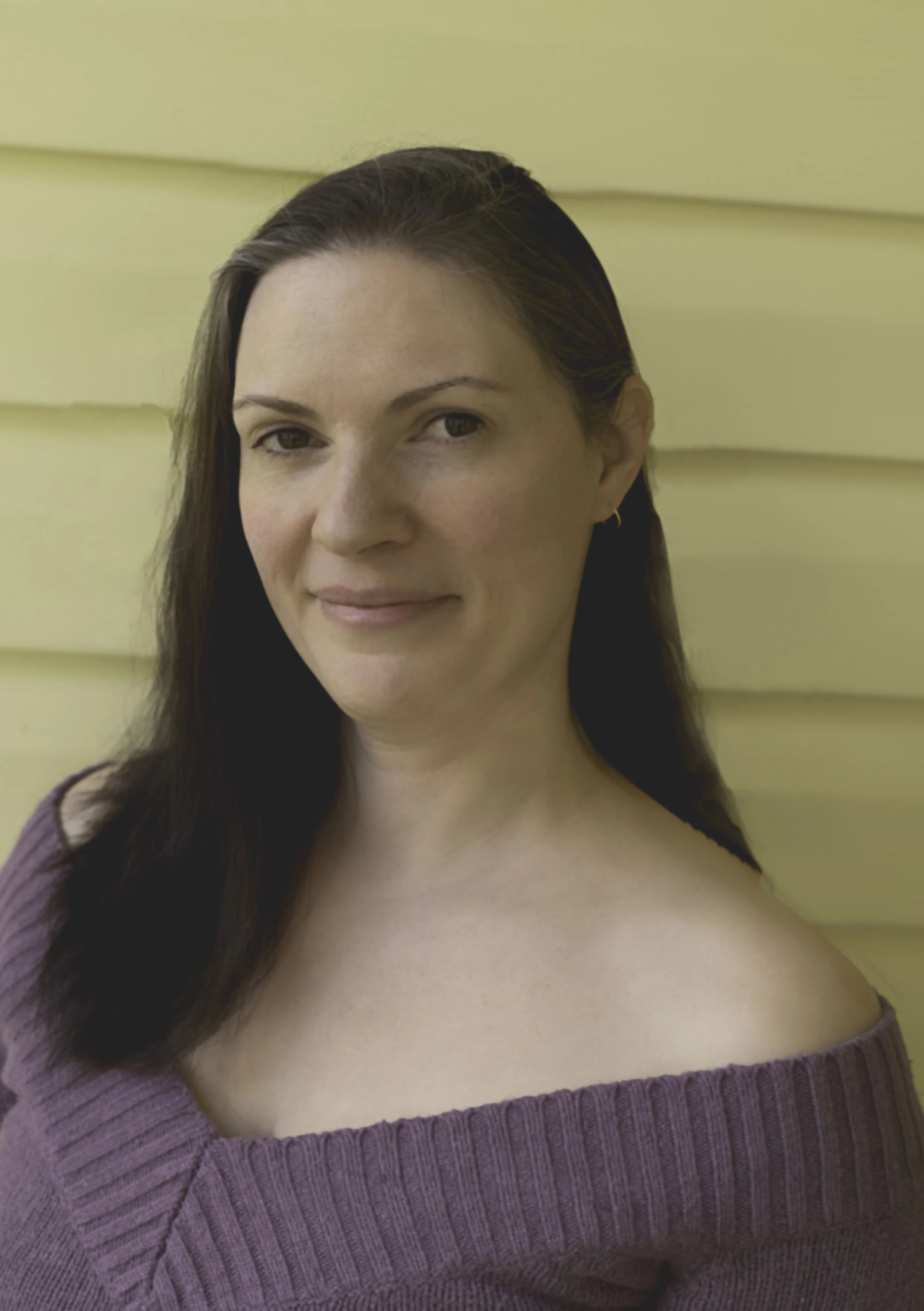 Portrait of a woman with long dark hair standing against a yellow wall.