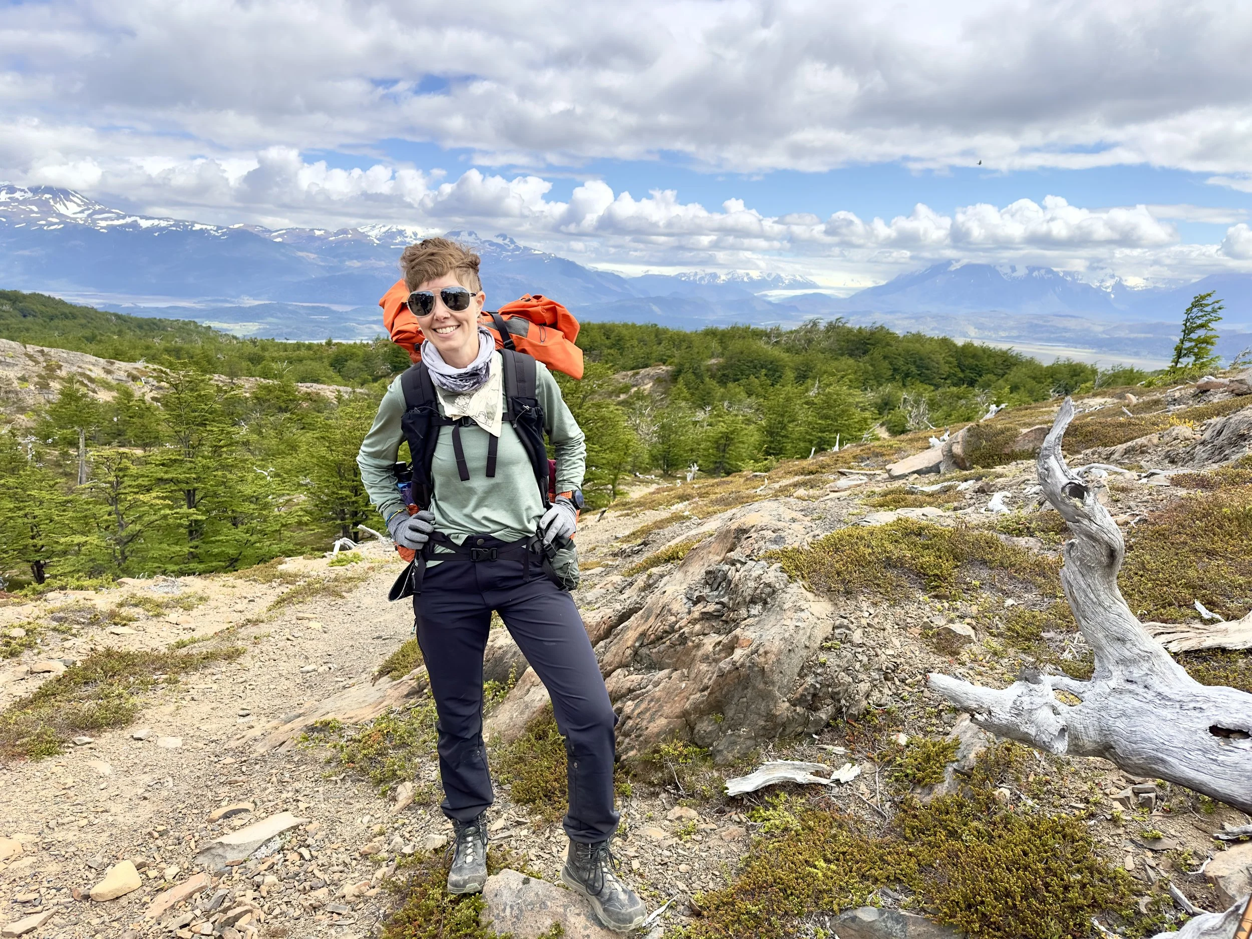A smiling woman dressed in outdoor hiking gear, standing on a rocky trail in a mountainous landscape with snow-capped peaks and clouds in the background.