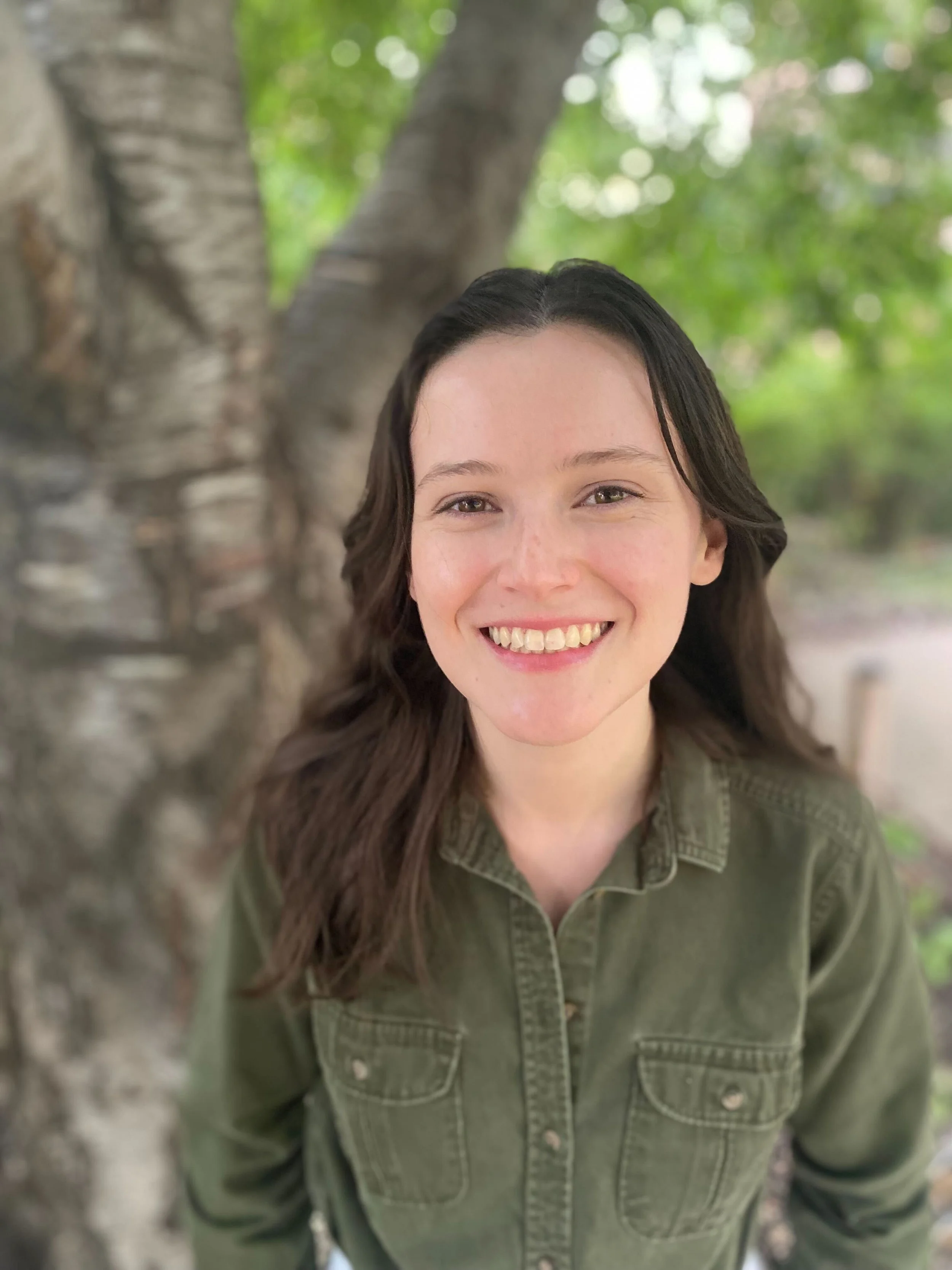 A smiling woman with dark hair and a green shirt standing outdoors near a tree with green leaves in the background.