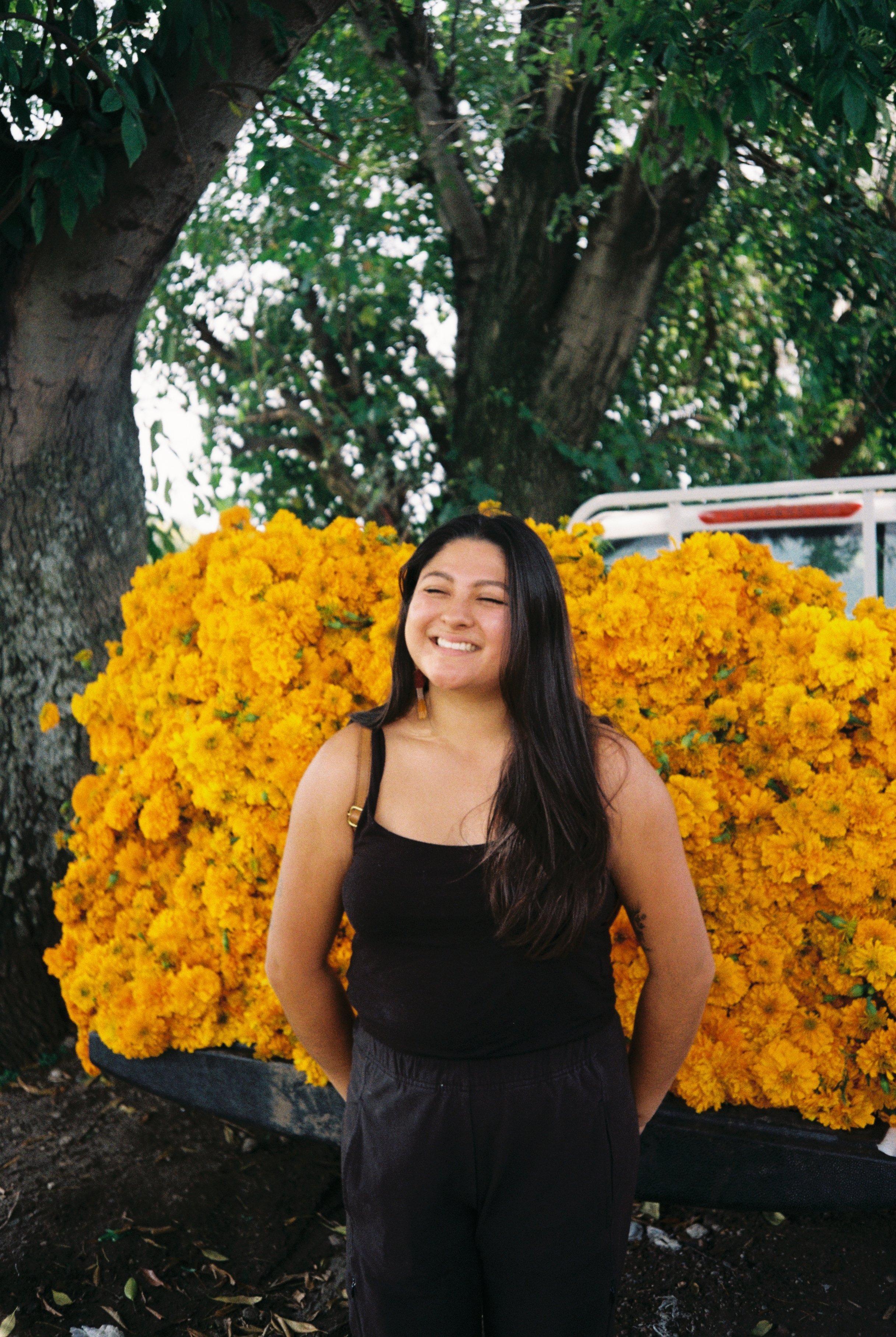 A young woman with long dark hair, smiling and standing outdoors in front of a large bush of vibrant yellow flowers, with trees in the background.
