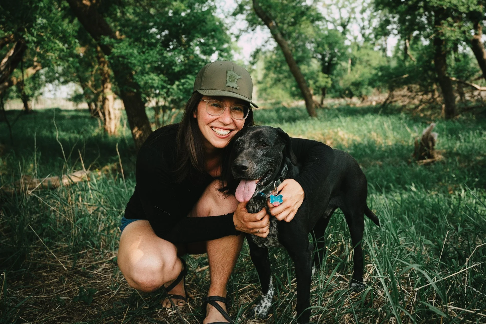 Woman crouching in a forest clearing, smiling, hugging a black dog with a blue collar and blue bone-shaped tag, surrounded by green trees and grass.