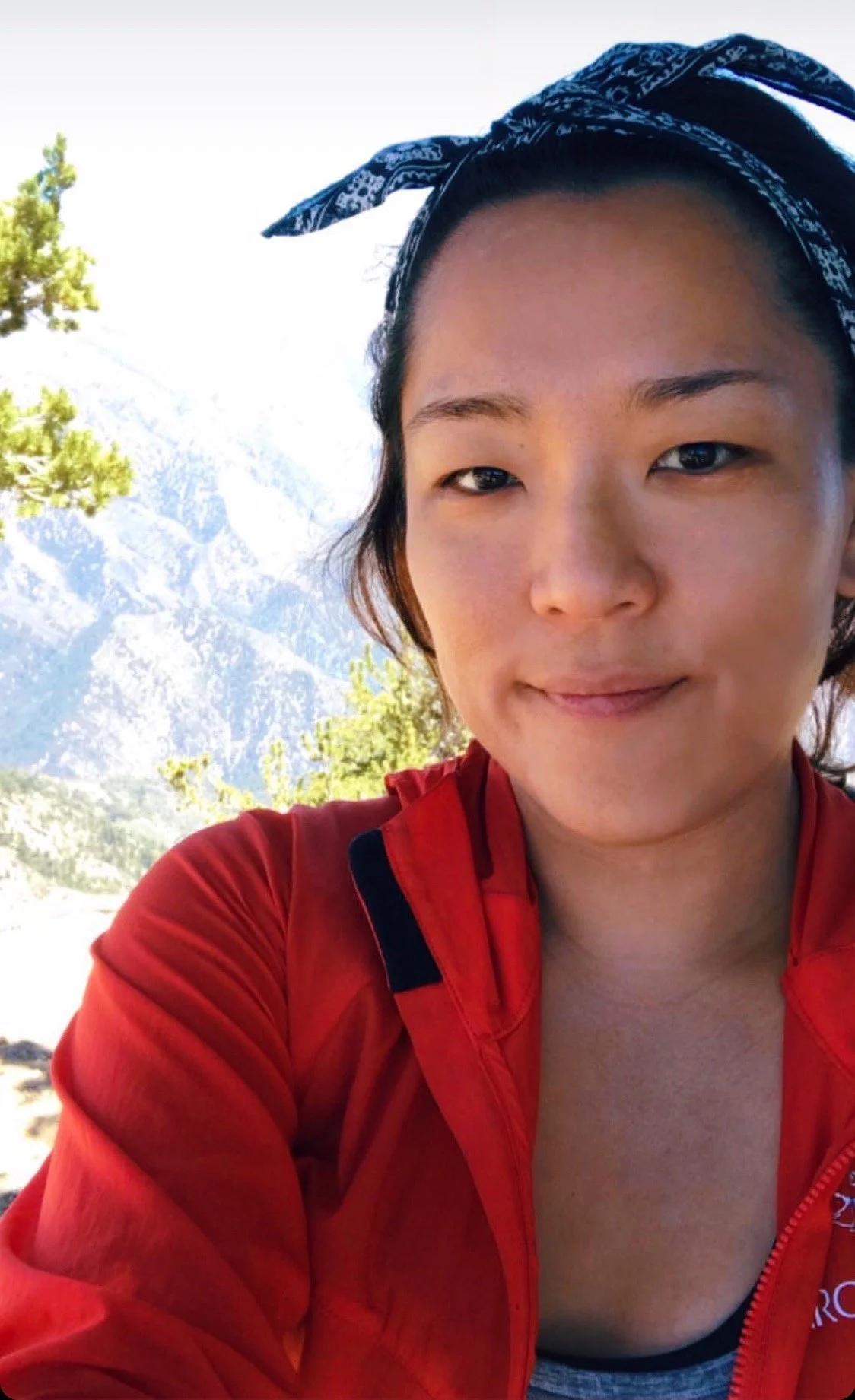A woman with a blue bandana on her head, wearing a red jacket, outdoors with trees and mountains in the background.