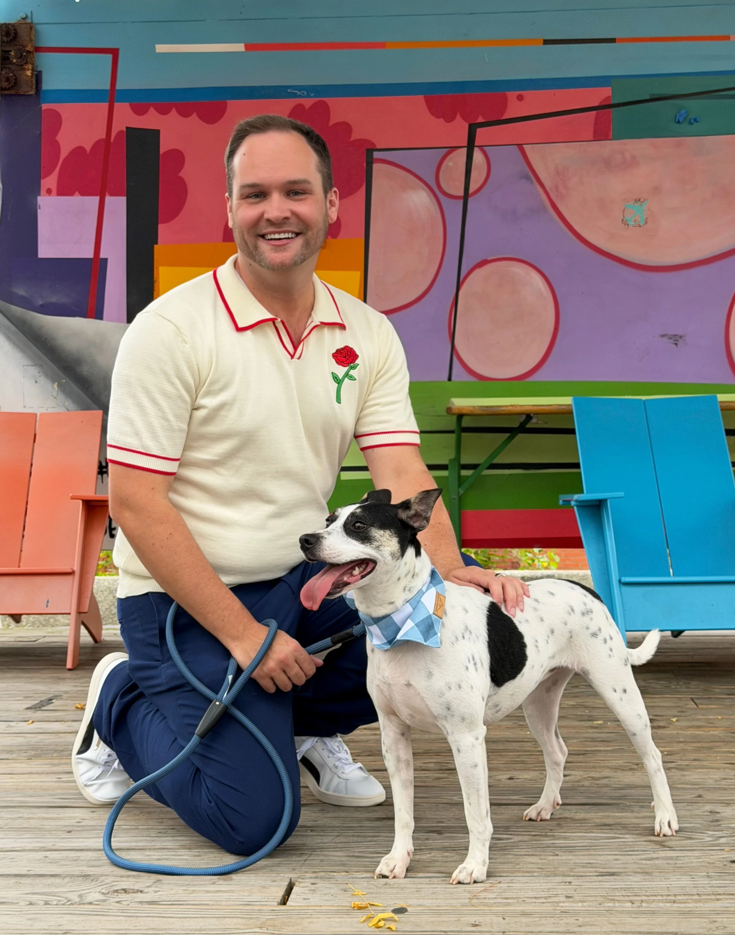 A man kneeling on a wooden deck next to a black and white dog with a blue bandana, both smiling, in front of a colorful mural with abstract shapes and bright colors.