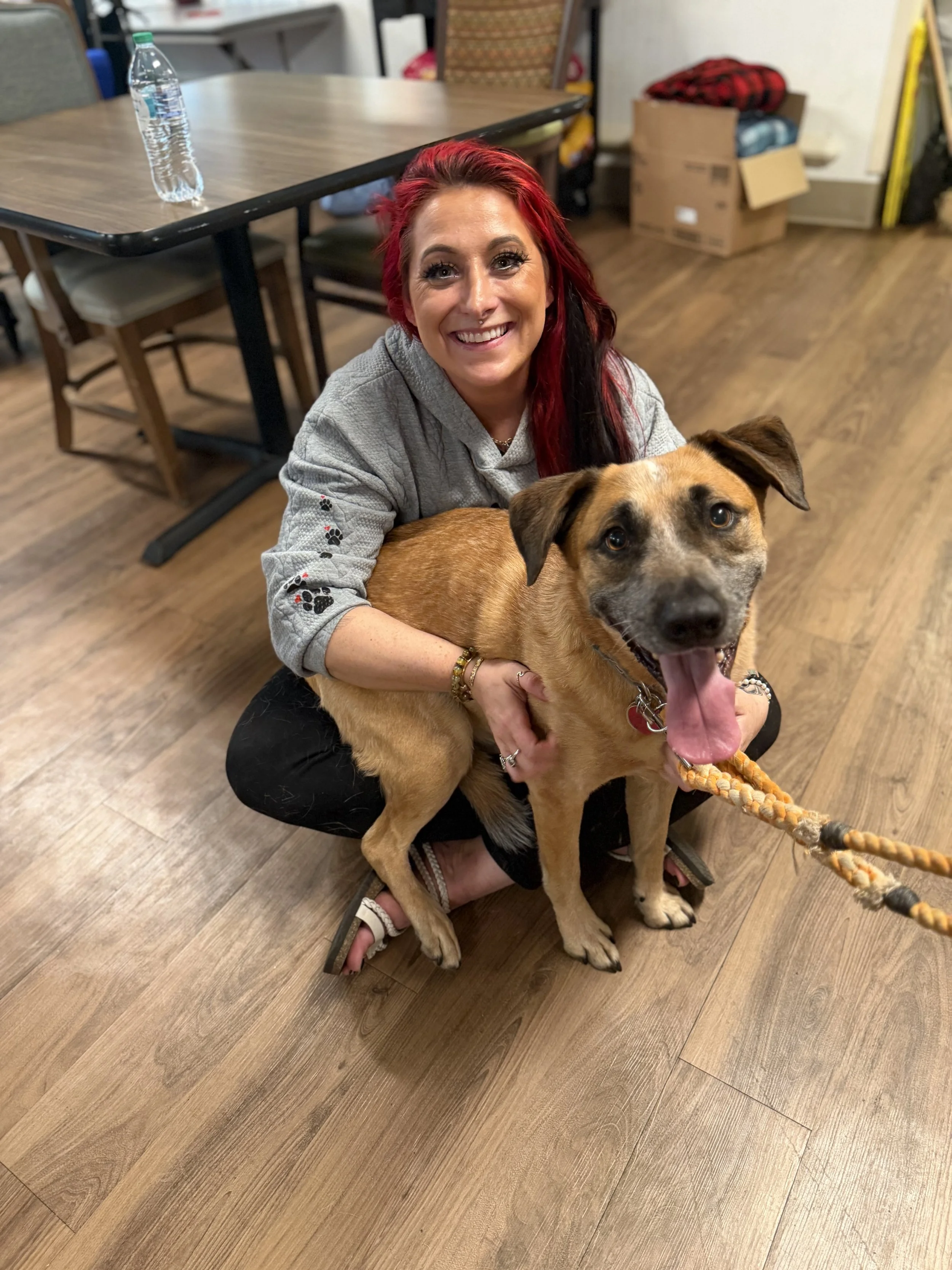 A woman with red and black hair smiling and holding a brown and black dog with a tan face indoors, with a table, chairs, and boxes in the background.