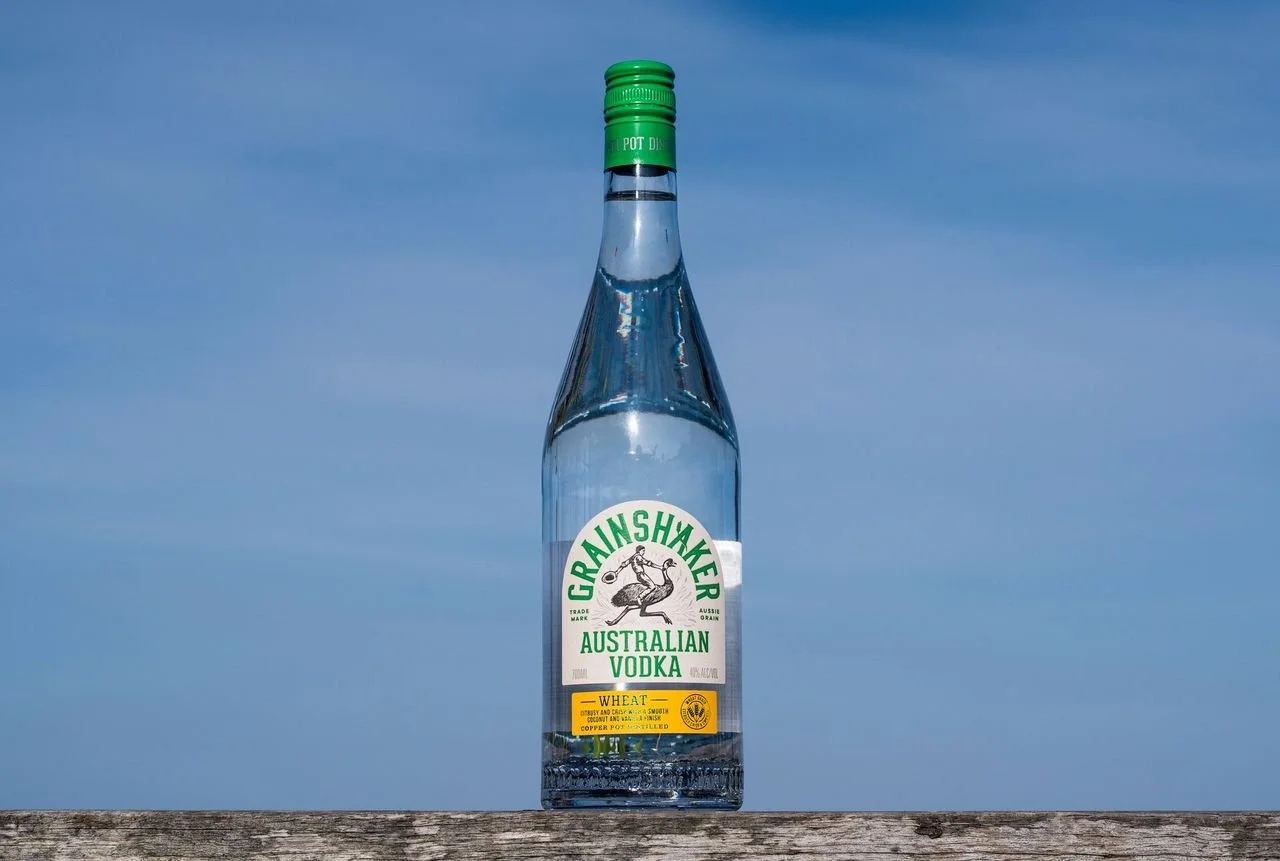 A clear glass bottle labeled 'Grainshaker Australian Vodka' with a green cap, resting on a wooden surface against a blue sky background.