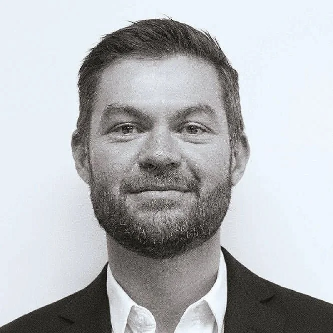 Black-and-white headshot of Greg Mitchell with a beard and short hair, wearing a suit jacket and white shirt, against a plain background.