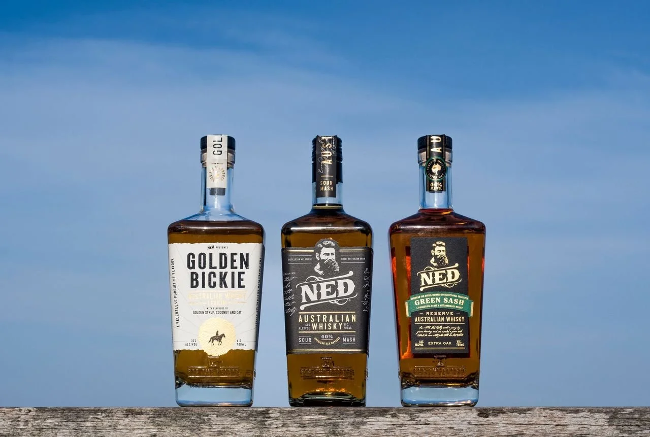 Three bottles of Australian whisky from Ned brand, placed on a wooden surface under a blue sky, featuring distinct labels and caps.