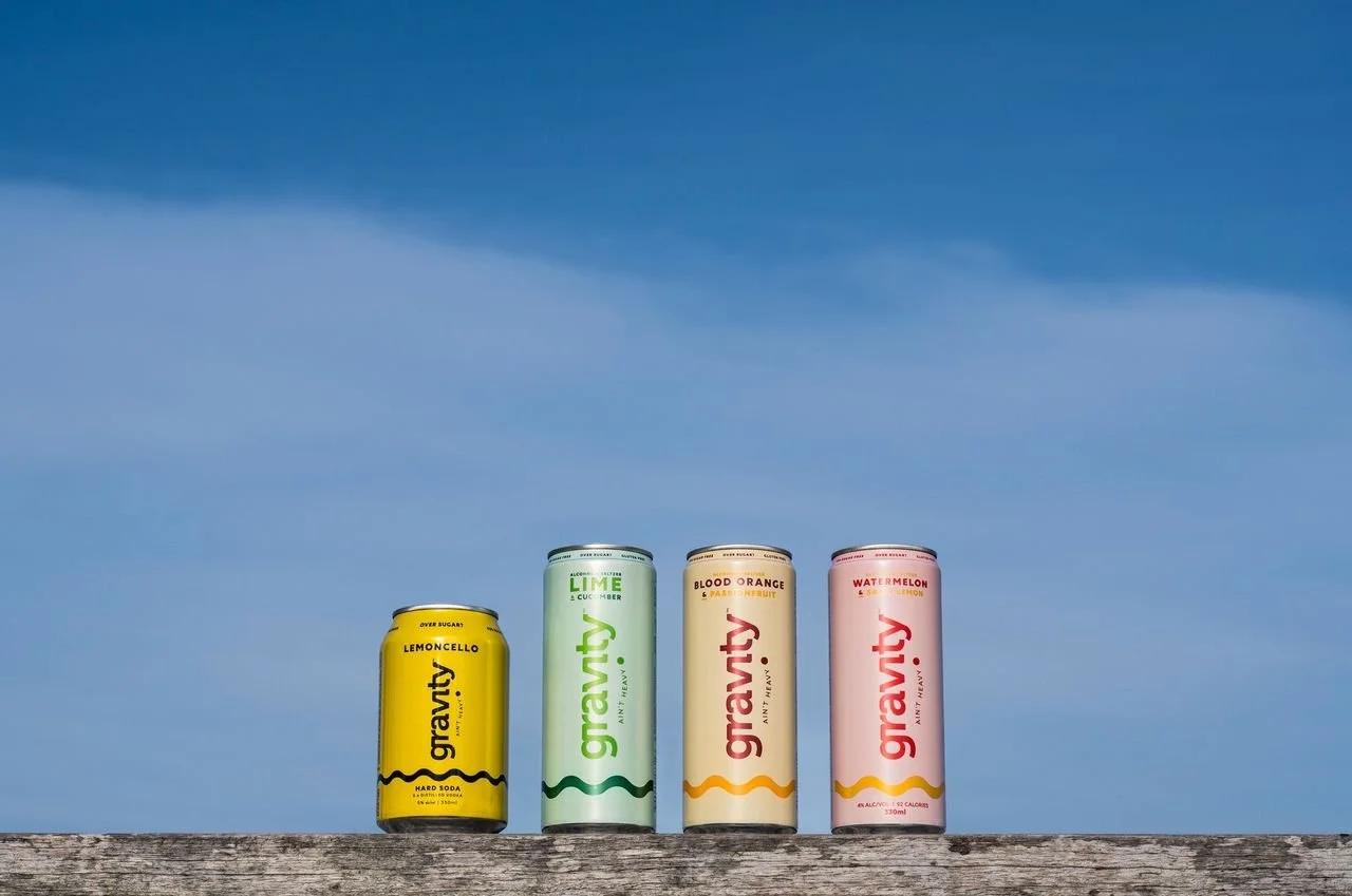 Four cans of Gravaty soda on a wooden surface against a blue sky, flavors from left to right: Lemoncello, Lime, Blood Orange, Watermelon.