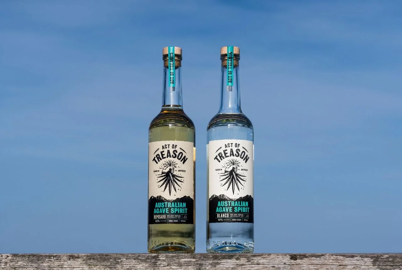 Two bottles of Treason Australian Grape Spirit, one Reposado and one Blanco, on a wooden surface outdoors against a clear blue sky.