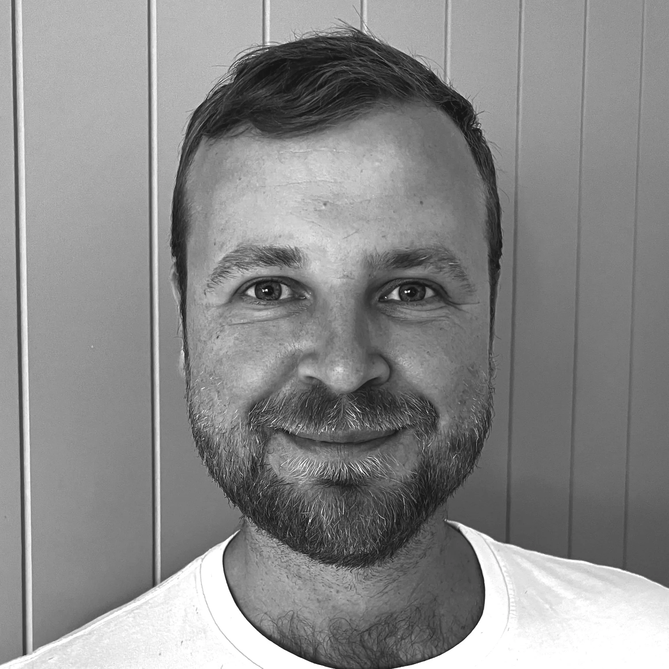 A black and white close-up portrait of a man with a beard and short hair, smiling, standing in front of wood paneling.