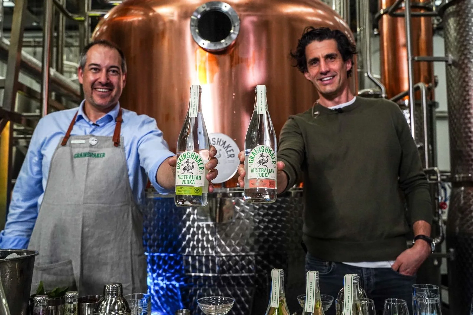 Two men smiling and holding bottles of Australian vodka in a distillery.