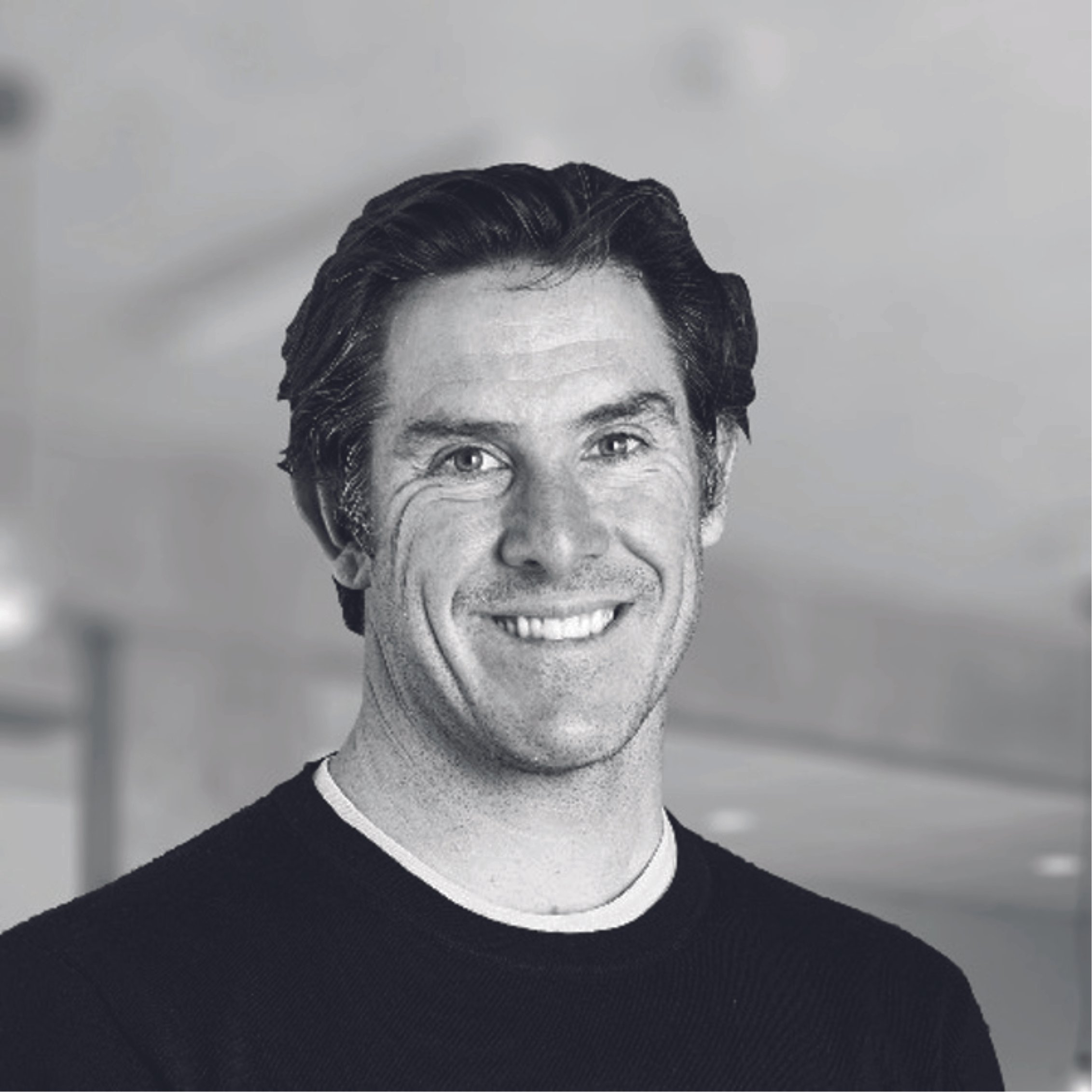 Black and white photo of a smiling man with dark, wavy hair, wearing a crewneck sweater with a light-colored collar.